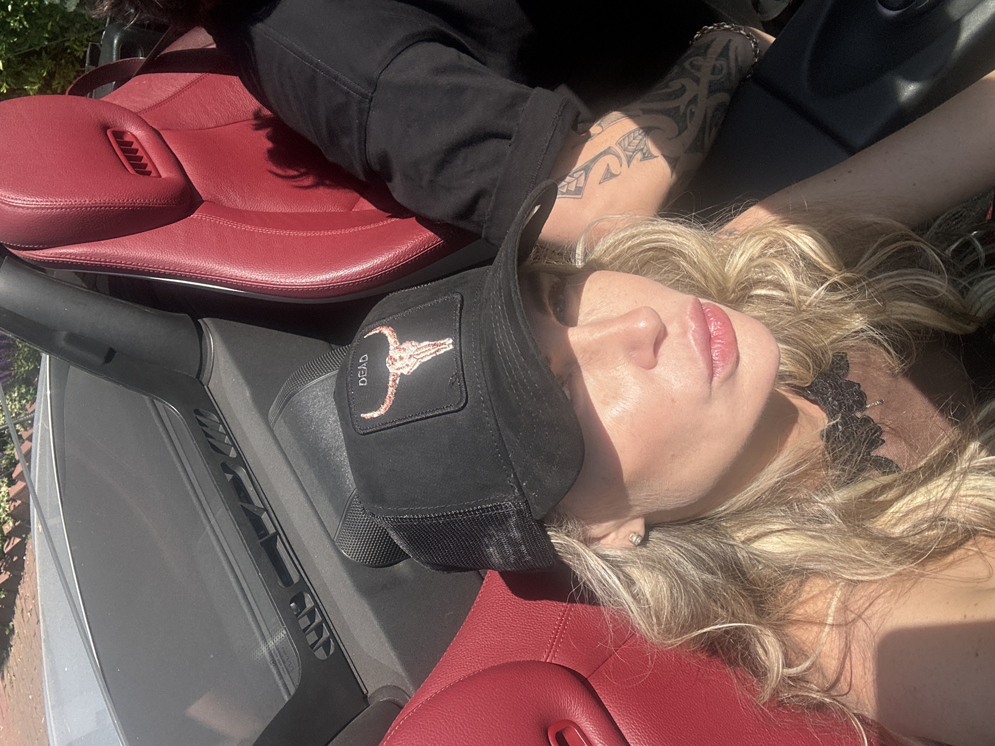 A woman with blonde hair is reclining in a convertible car with red leather seats. She wears a black baseball cap with an emblem featuring a bull skull and the word "DEAD." The woman has on a black lace necklace and an earring. Her left arm has a tattoo. The car's interior and exterior are visible, including the dashboard and what appears to be a glimpse of foliage outside the car. There is no specific indication of the city of Katwijk, Netherlands in the image.