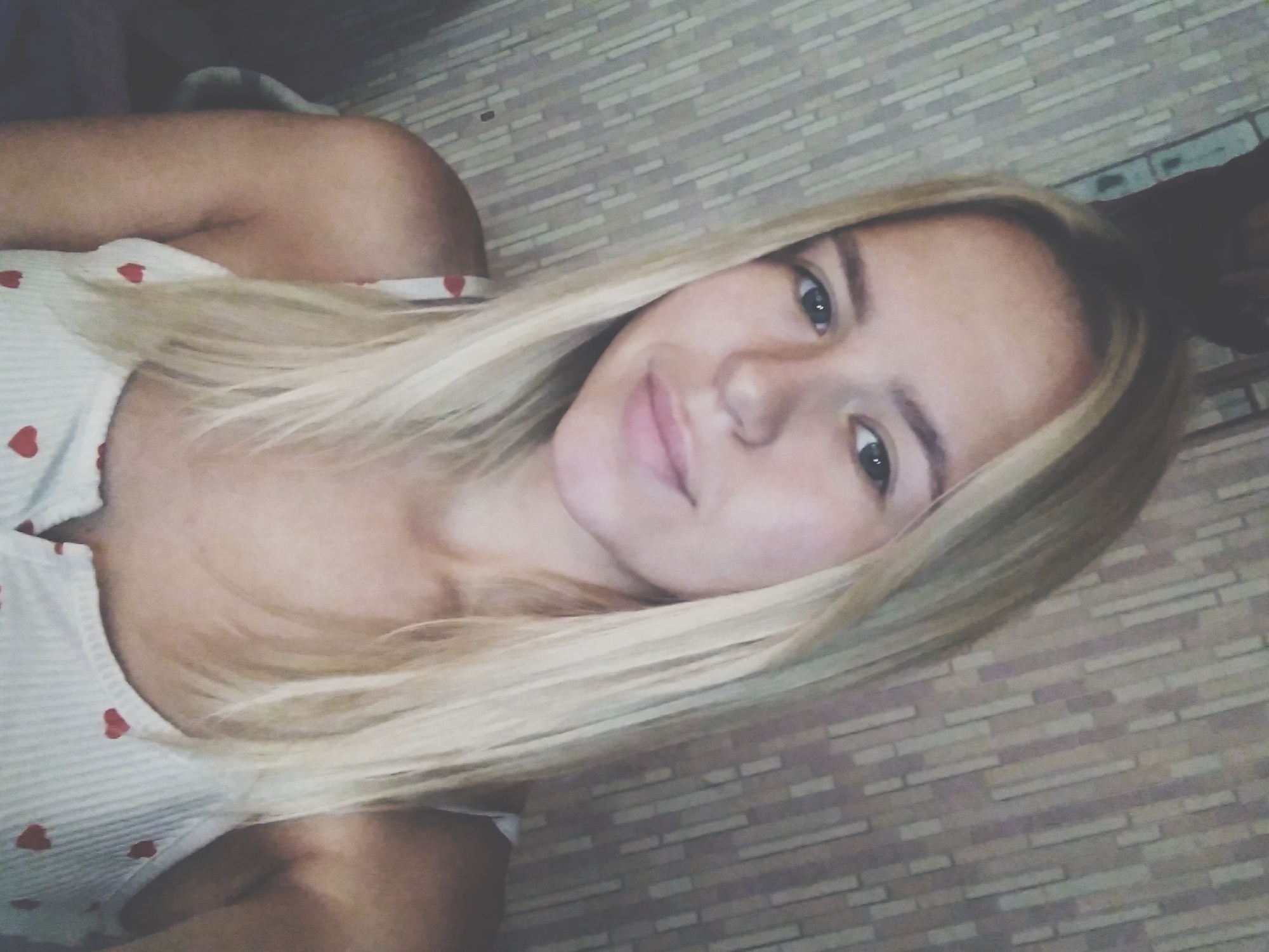 The image is a selfie taken by a young woman with blonde hair. She is wearing a white camisole with small red hearts. The background appears to be a wall with a textured, striped pattern. The image quality is somewhat blurred and grainy, with a sepia-toned filter applied. The woman's face is the primary focus, and she is looking directly at the camera. No specific location within the city of Radisne, Ukraine, can be identified from the visual information provided.
