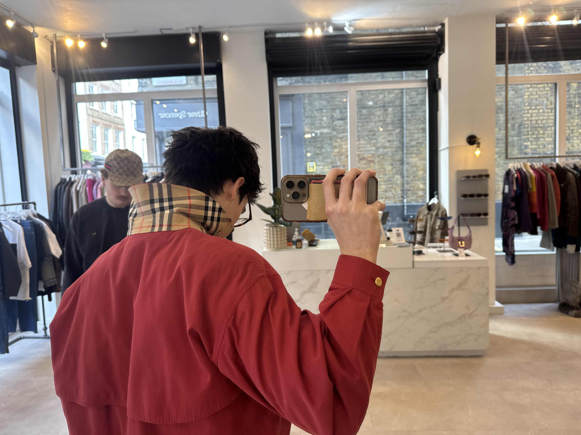 A person wearing a red jacket with a tan and black checkered collar is taking a photo with their phone in a clothing store. A second person, wearing a hat, is standing behind them, looking down. Clothes are visible on racks in the background. Outside, there is a view of buildings in London, United Kingdom.