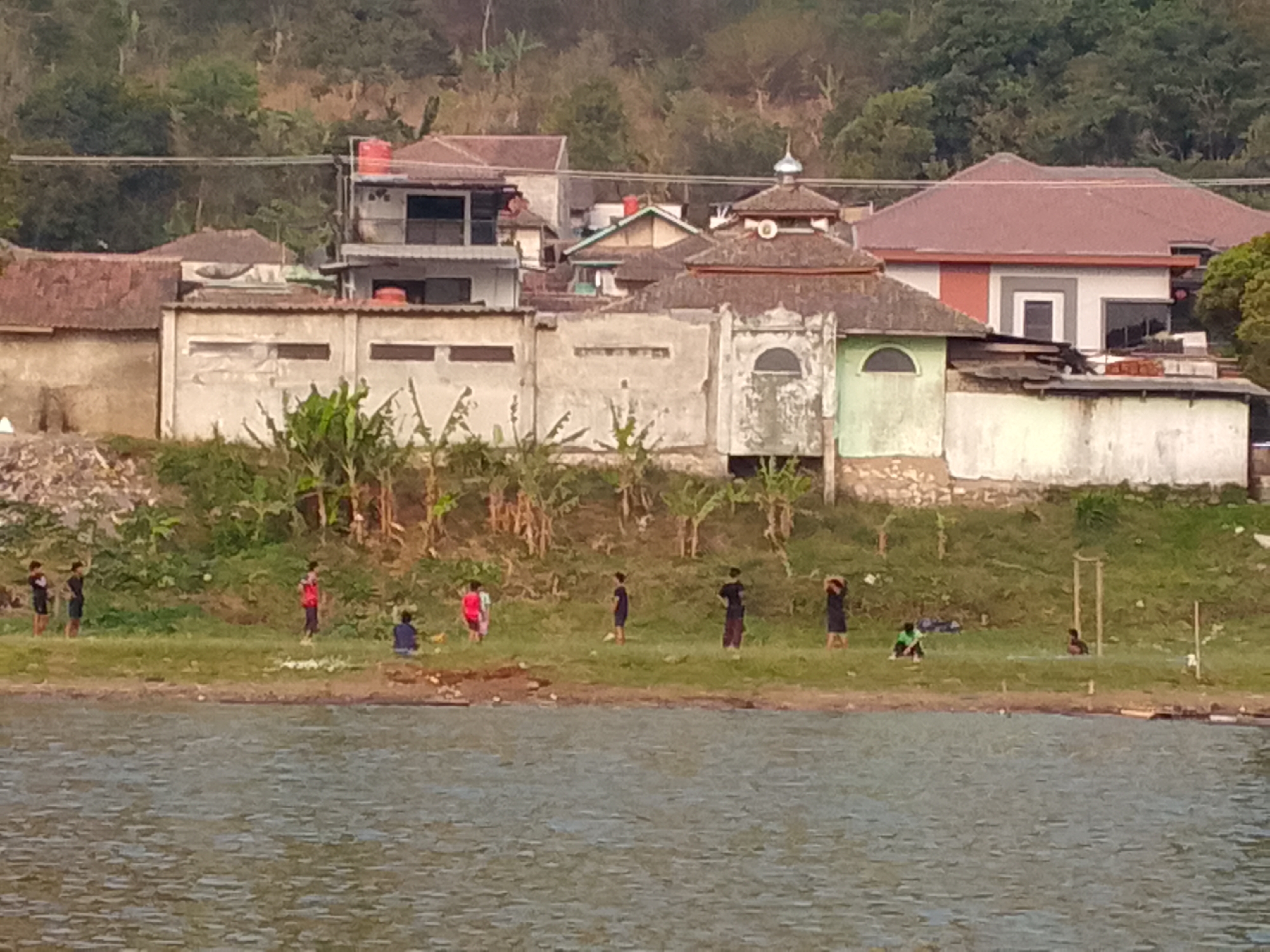 The image depicts a lakeside scene in Ciburuy, Indonesia, with a cluster of buildings situated on a grassy embankment overlooking a body of water. The buildings appear to be a mix of residential structures and what may be a mosque, characterized by a dome. Several people, mostly children, are visible on the grassy area near the water's edge. Some are standing, while others are sitting or appear to be engaged in activities. The water itself is rippled, suggesting a slight breeze or movement. The background shows a hillside covered with vegetation and trees.