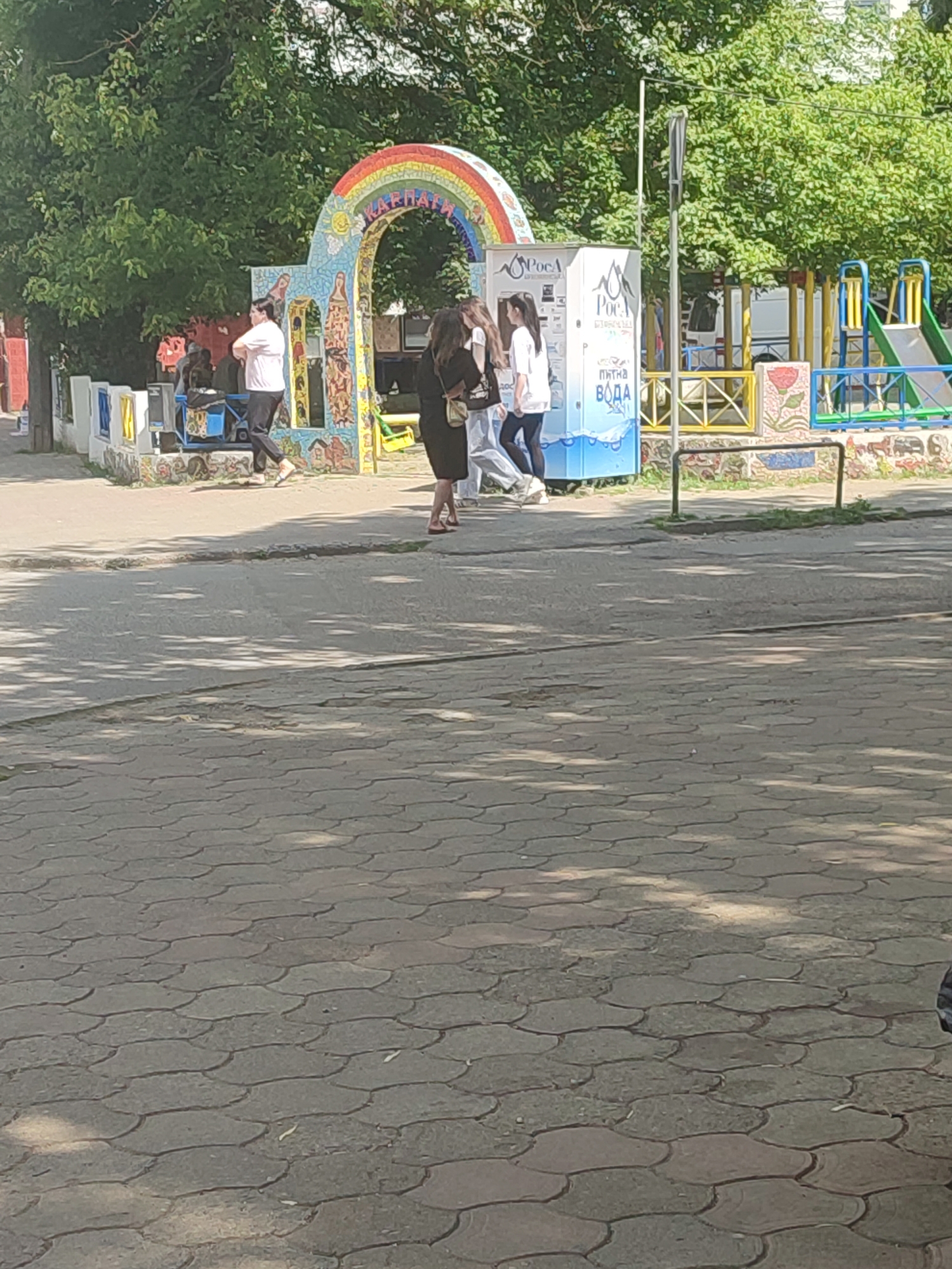 A group of people are gathered near a colorful mosaic archway with a rainbow on it. The archway is decorated with various images and patterns. To the right of the archway is a vending machine with Cyrillic text and images of water bottles. Beyond that, a children's playground with slides and colorful fencing is visible. A person wearing a white t-shirt and dark pants is standing near a blue object under the archway. Several other individuals, including two women in casual attire, are in the vicinity. The scene is set outdoors, with trees providing shade and a paved ground surface in the foreground. This location appears to be a public area in the city of Chernivtsi, Ukraine, possibly a park or a recreational space.