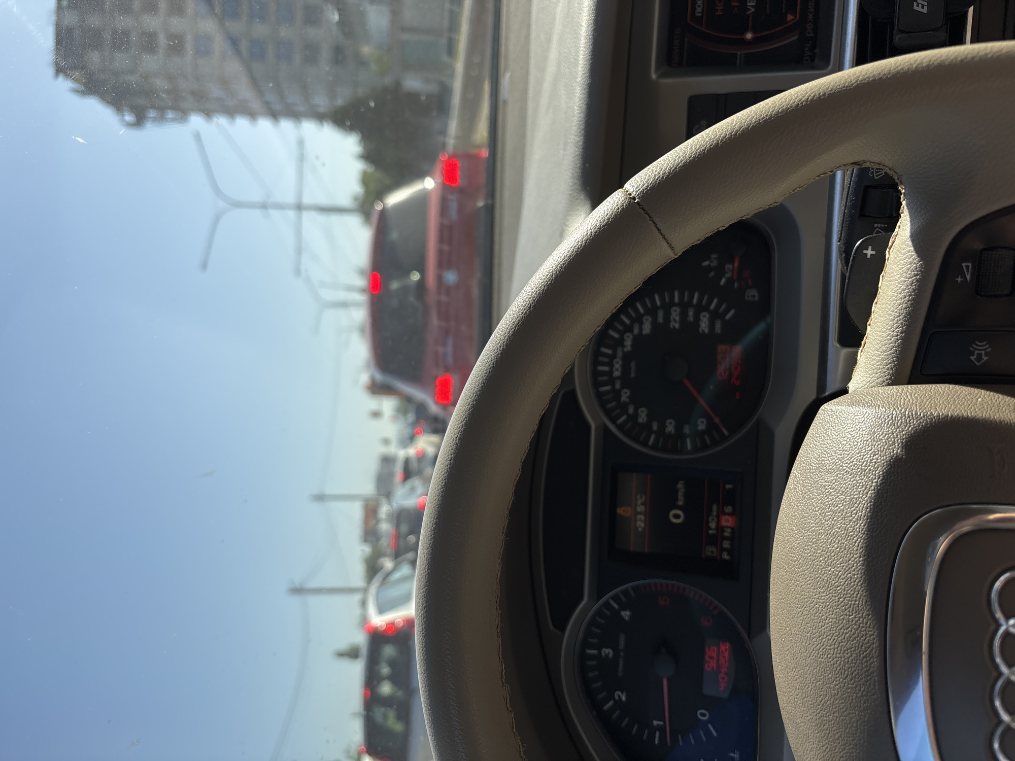 The image shows the interior of a car, looking through the windshield at a city street with heavy traffic. In the foreground is the steering wheel, dashboard, and instrument cluster of the car. The steering wheel is made of beige leather and has some stitching detail. The instrument cluster includes a speedometer, tachometer, and a display showing temperature (-23.5°C), speed (0 km/h), and gear selection (P R N D S 1). Outside the car, several vehicles are visible, including a red car in front of the camera car. A tall, unfinished building is visible on the right side of the frame, along with streetlights and trees. The sky is clear and blue. The scene appears to be captured during daylight hours.