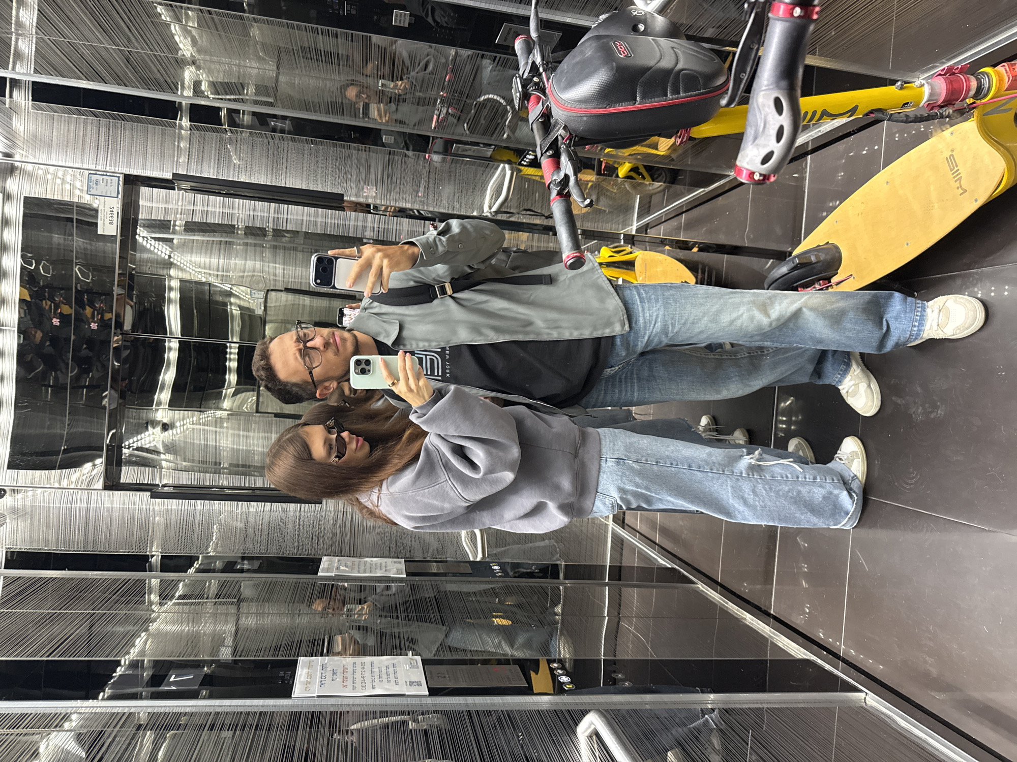 Inside a modern elevator, two individuals, likely a couple, are taking a selfie. A man with glasses is holding up a smartphone, while a woman with sunglasses also holds her phone up to participate. Behind them is a yellow electric scooter, suggesting their location may be a public transit hub or residential building. The elevator interior features a metallic design with mirrored surfaces, creating reflections of the people, scooter, and surroundings. The walls have a textured appearance. Given the context of previous queries and the location context, the scene is most likely taking place somewhere in the city of Tel-Aviv, Israel.