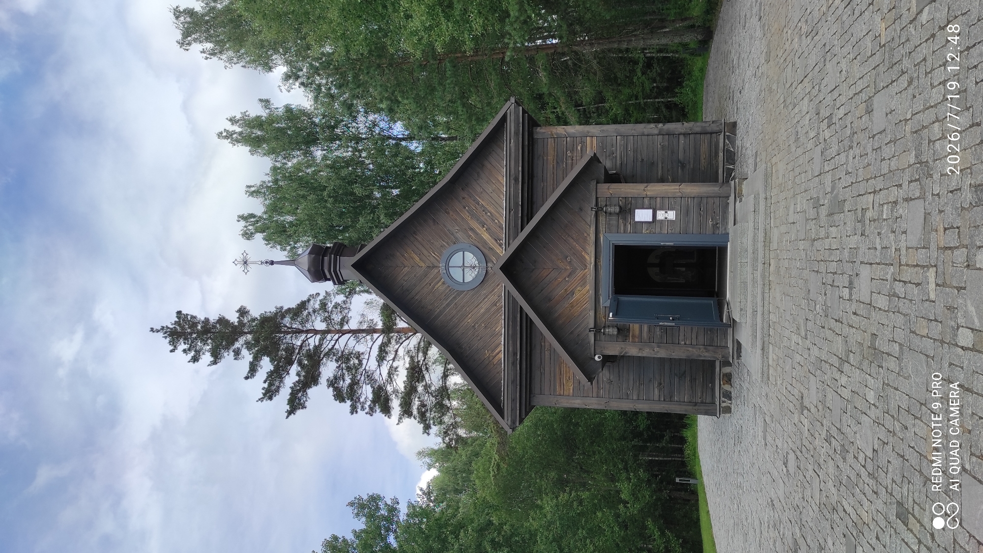 A rustic, dark wooden chapel stands centrally in the frame, its gabled roof adorned with a small, dark dome topped with a cross. The chapel features a central entrance with an open, dark blue door revealing a dimly lit interior, flanked by two outdoor lamps. Above the entrance, a small circular window with ornate detailing is visible. The chapel is set against a backdrop of lush green trees, with a tall pine tree to the left and other deciduous trees to the right. The ground in front of the chapel is paved with grey cobblestones, forming a walkway leading to the entrance. The sky above is partly cloudy with a mix of blue and grey clouds. The image appears to be taken on July 19, 2026, at 12:48 PM, in Каменскі сельскі Савет, Belarus.