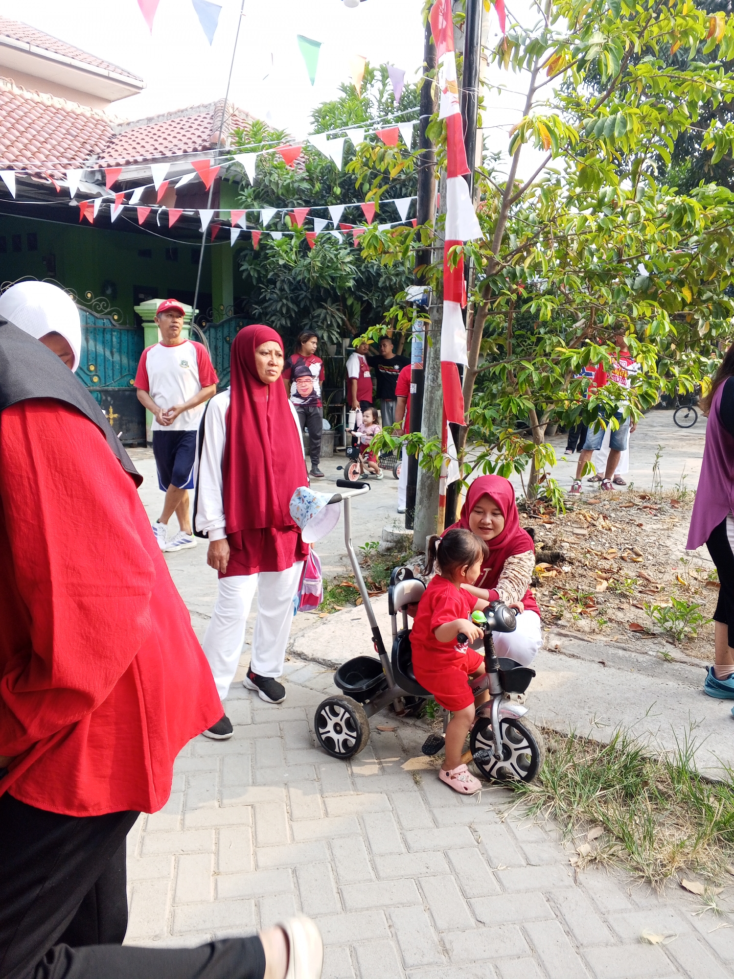 The image shows a group of people outdoors, likely in a residential area, celebrating a national holiday given the red and white flags and bunting visible. In the foreground, a woman wearing a red hijab and white clothing is crouched down, smiling at a young child in a red shirt who is sitting on a tricycle. The child is holding onto the handlebars, and their feet are on the pedals. Behind them, other individuals are standing and walking. To the left, a person in a red outfit and white head covering is partially visible. In the background, there are houses with red-tiled roofs and lush green trees. A man in a red and white shirt and a red cap is standing, and other people in similar attire are seen further back. The ground is paved with interlocking bricks. The scene appears to be in Ciruas, Indonesia, as indicated by the context.