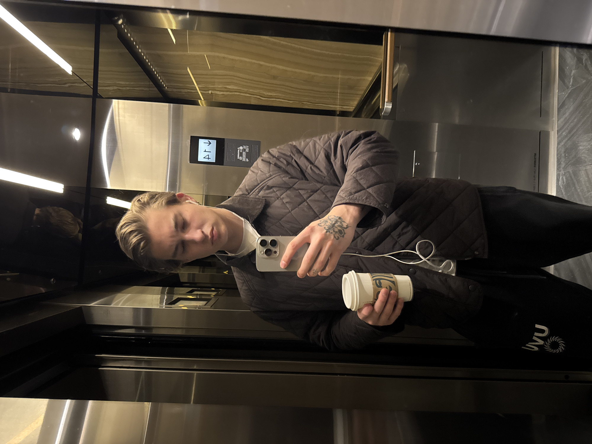 A young man is taking a selfie in an elevator. He is wearing a quilted jacket and black pants with the letters "UWU" printed on them. He is holding a cup of coffee in one hand and a smartphone in the other. The elevator has stainless steel walls and a control panel with buttons for different floors. Based on the interior design of the elevator, including its lighting and paneling, and the style of the elevators themselves, this may be taken in a modern building located in London, United Kingdom.