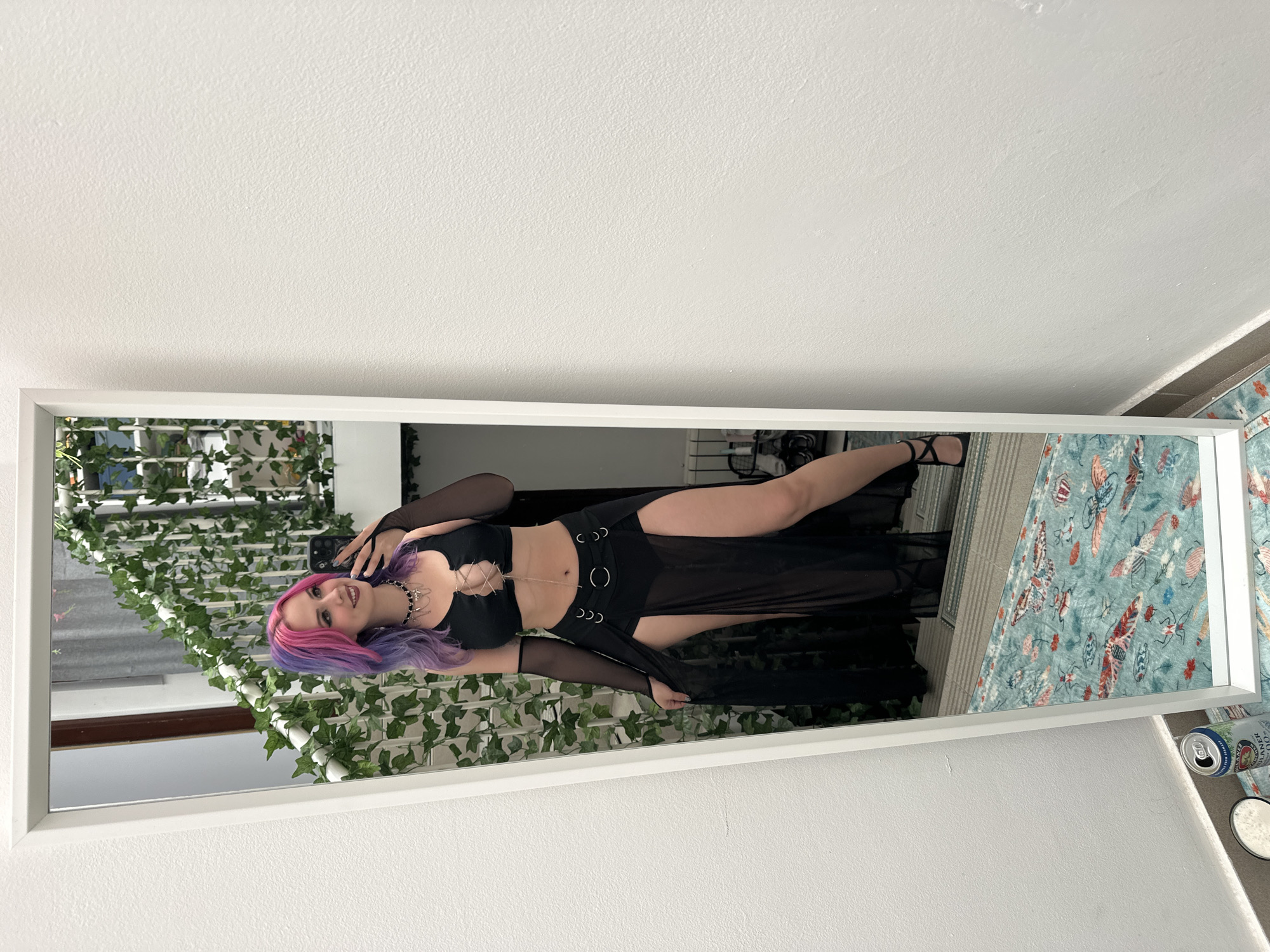 A woman with purple and pink hair is standing in front of a full-length mirror, taking a selfie with her phone. She is wearing a black outfit consisting of a top with a laced chest and a skirt with attached metal rings. The mirror is framed in white and has artificial vines draped along the top left side. In the reflection, there is a blue and green patterned bedspread, a window with gray curtains, and a glimpse of a room. Two cans and a small round container are visible on a surface to the right of the mirror. The photo appears to be taken indoors against a white wall. The location is not clear enough to be confirmed.
