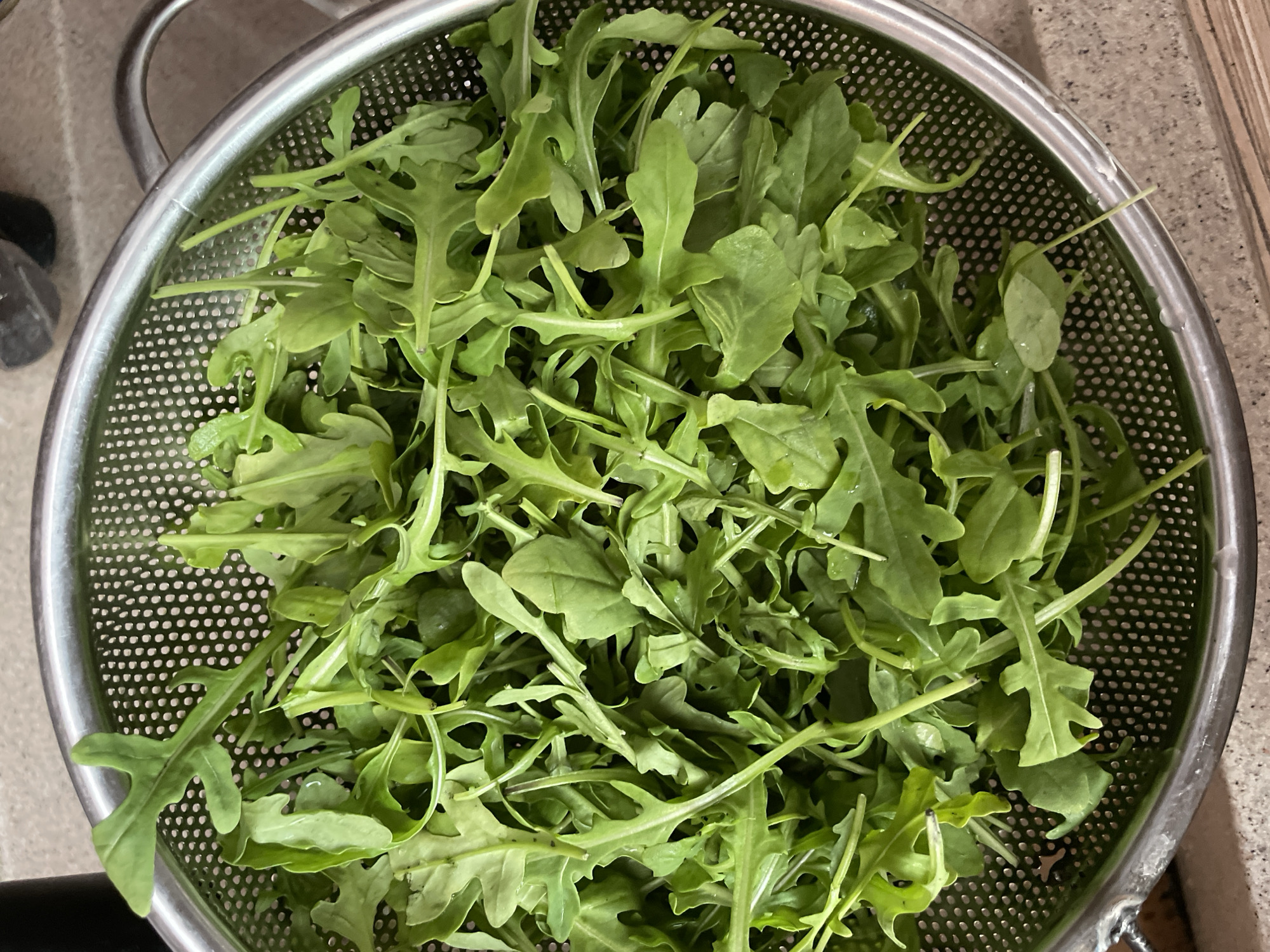 The image shows a close-up, overhead view of fresh arugula in a stainless steel colander. The arugula leaves are vibrant green and appear slightly damp, with some water droplets visible on the leaves and in the colander. The colander has a fine mesh with many small perforations and a rim of polished metal. The background is out of focus, but appears to be a countertop surface in muted tones. There are no people or animals visible in the image. The scene is indoors, likely in a kitchen, given the presence of a colander and food preparation elements. There is no information within the image that would allow for its location to be identified in the city of Zheleznogorsk, Russia.