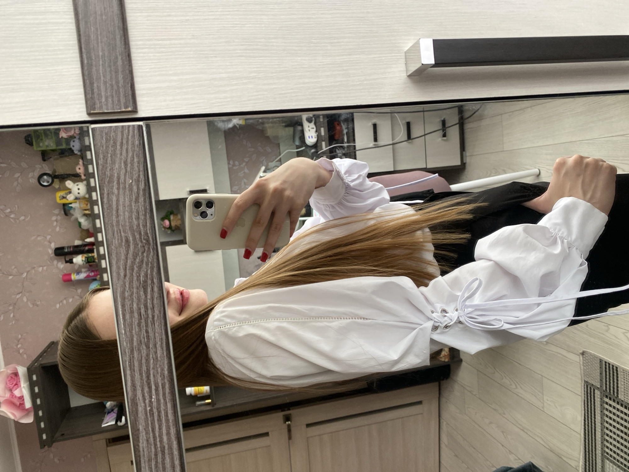 A young woman with long brown hair is taking a mirror selfie in a room. She is wearing a white blouse with puff sleeves and a black skirt. Her face is partially obscured by the mirror's frame. In the background, a closet with shelves and various items, including decorative objects, can be seen. The floor is wooden, and a rug is visible. The image appears to have been taken indoors, possibly in a bedroom or dressing area. No specific location within the city of Дьяково, Russia, can be identified from the image.