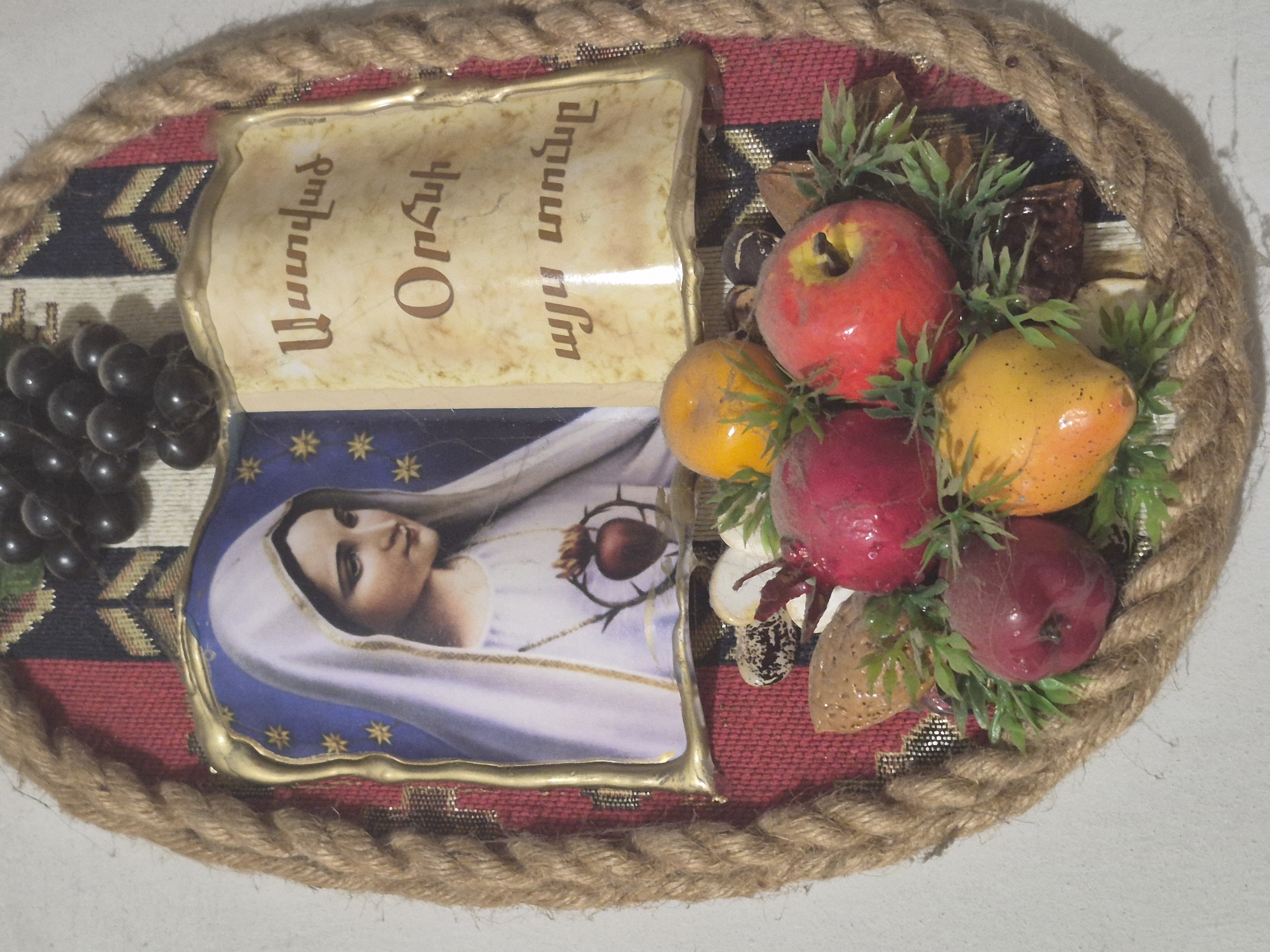 An oval-shaped, decorative plaque is pictured, likely displayed in a home in Vanadzor, Armenia. The plaque features a religious icon of Mary, the mother of Jesus, depicted with a white veil and a Sacred Heart symbol. To the right of the icon, there is text in Armenian script. At the bottom of the plaque, a cluster of artificial fruits, including red apples and yellow oranges, along with greenery, is arranged. The entire piece is framed by a thick, braided rope. The background appears to be a wall.