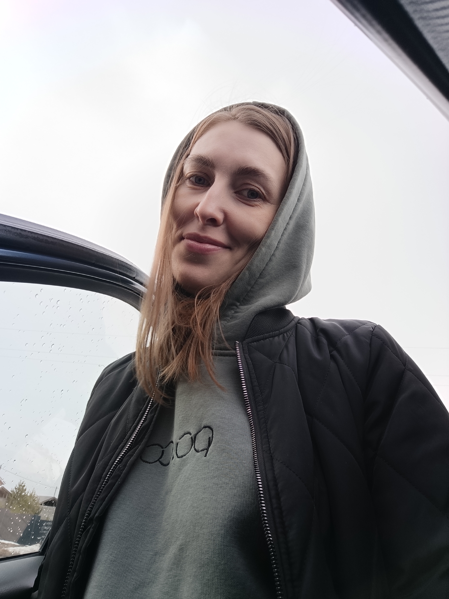 A woman in a black quilted jacket over a grey hoodie stands near a car. The car window is partially visible and has raindrops on it. Outside the car, there is a building, possibly a house, and a few trees. Given the information in the prompt, the location is Chaikovsky, Russia.