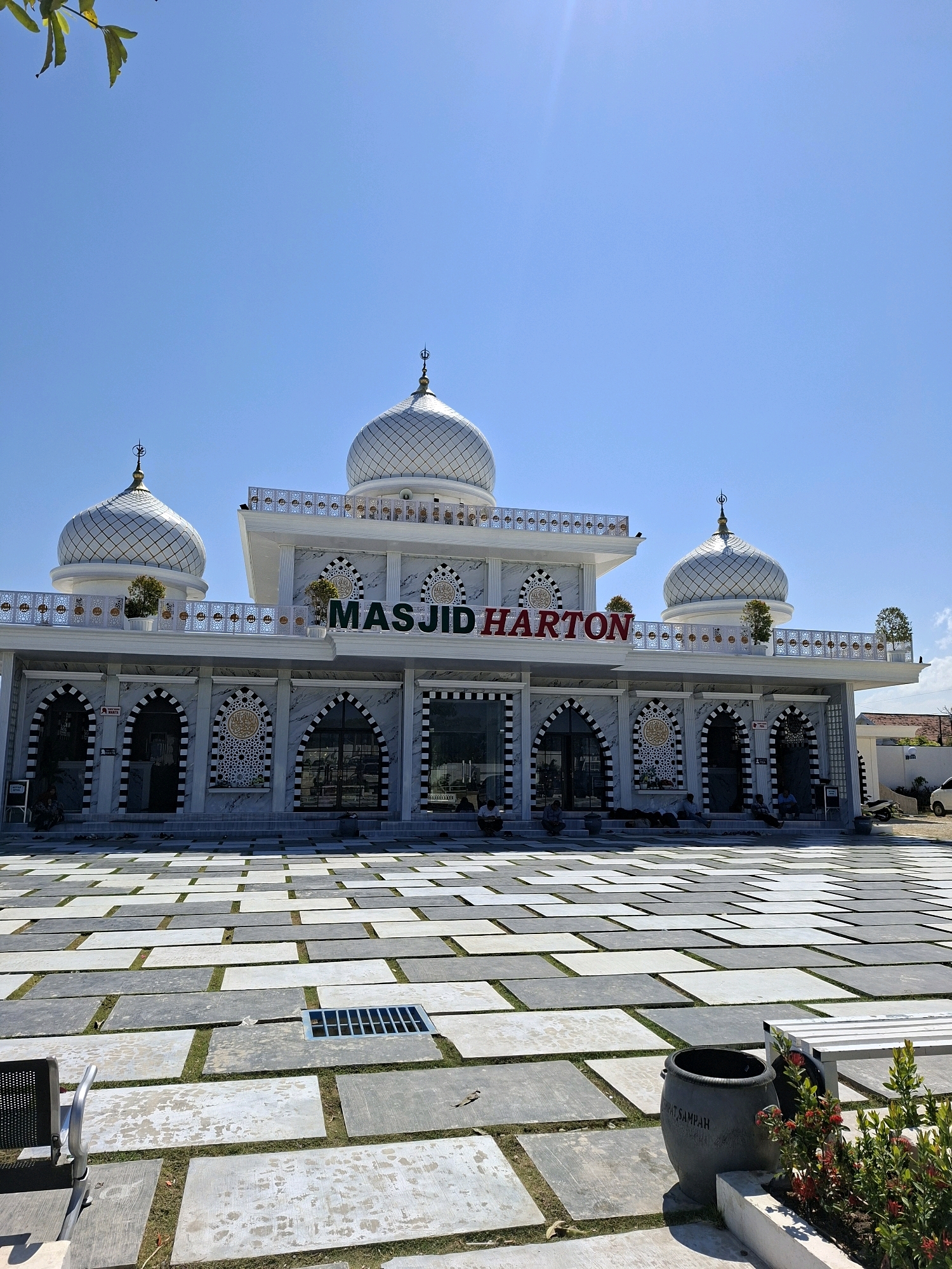 The image shows the Masjid Harton mosque, located in Camplong, Indonesia. The mosque features a white facade with distinctive black and white striped arches and decorative panels. It has multiple domes topped with crescent moons, typical of Islamic architecture. Large stone tiles form a courtyard in front of the mosque, with some grass growing between them. Several people are seated or standing in the courtyard and on the steps leading to the mosque entrance. In the foreground, a large dark-colored trash bin with Indonesian writing is visible, along with some red flowers and a bench. The sky is clear and blue.