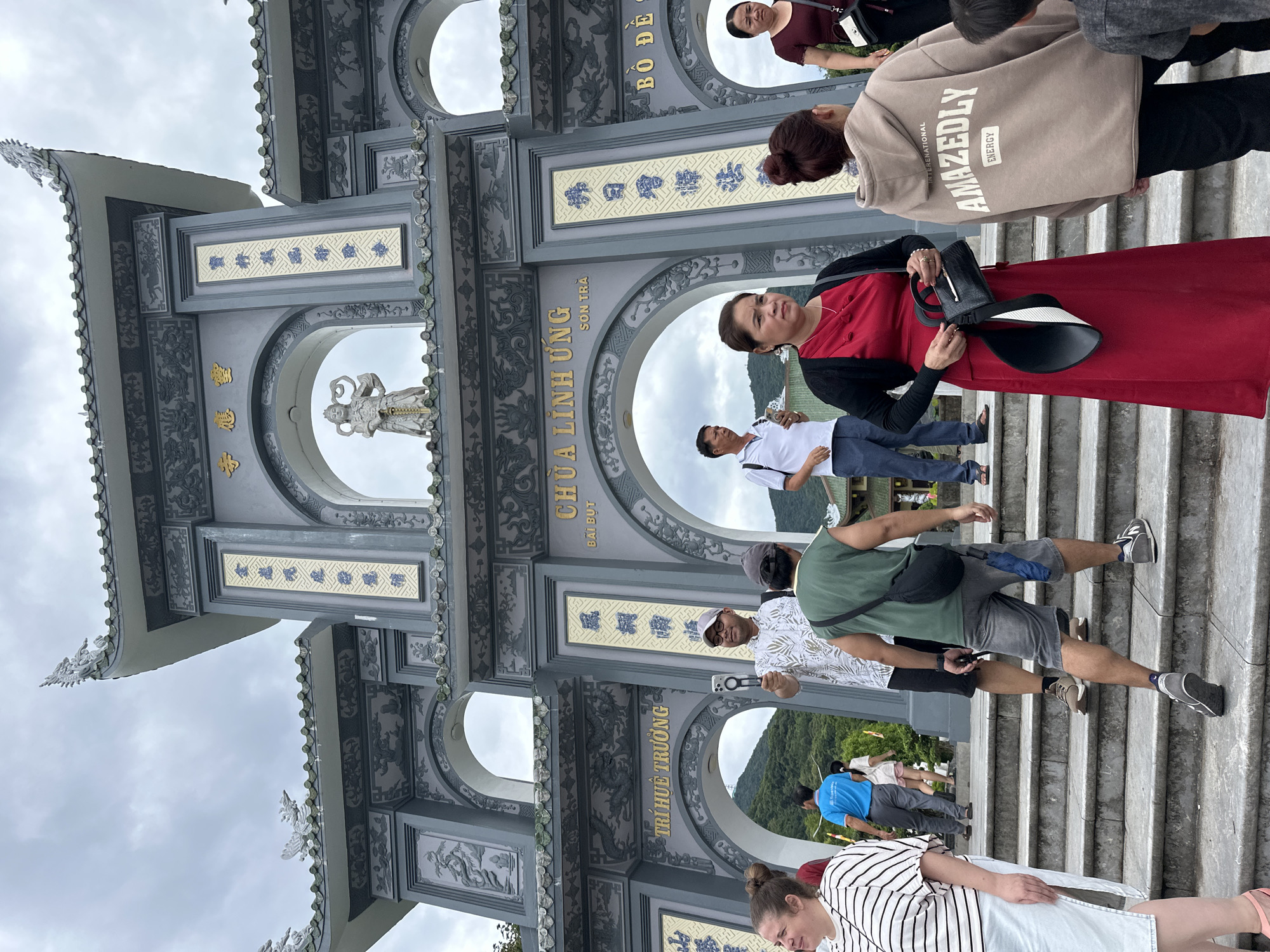 The image shows the entrance to Linh Ung Pagoda on Son Tra Peninsula in Da Nang, Vietnam. A group of tourists is ascending a wide stone staircase leading up to the ornate, grey archway of the pagoda. The archway is decorated with intricate carvings of dragons and other motifs, and features vertical panels with Vietnamese inscriptions. A statue of a deity is visible in the upper arch. The sky is overcast. On the stairs, people are dressed in casual summer attire. In the foreground, a woman in a striped shirt and white skirt walks past. Behind her, a man in a patterned shirt is holding up a phone, appearing to record or take a photo. Further up, a man in a green sleeveless shirt walks with a black bag slung over his shoulder. To the right, a woman in a red dress stands holding a handbag and a wide-brimmed hat. Another woman in a tan hooded sweatshirt with the words "AMAZEDLY ENERGY" is also in the foreground. In the background, lush green mountains are visible, partially obscured by the pagoda structure.