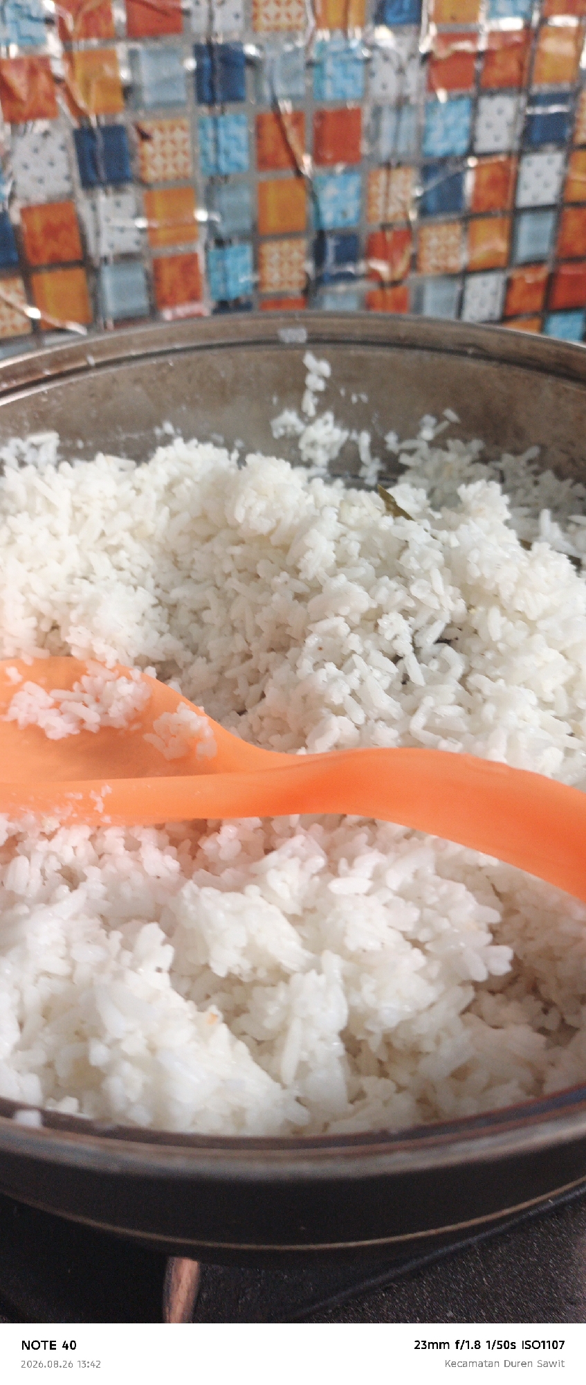 The image shows a close-up of a pot filled with white rice, with an orange rice paddle resting on top of it. The background features a colorful tiled wall. At the bottom of the image, there is text that reads "NOTE 40 2026.08.26 13:42 23mm f/1.8 1/50s ISO1107 Kecamatan Duren Sawit". This indicates the photo was taken in Kecamatan Duren Sawit, a subdistrict within East Jakarta, Indonesia, on August 26, 2026, at 13:42, with specific camera settings.