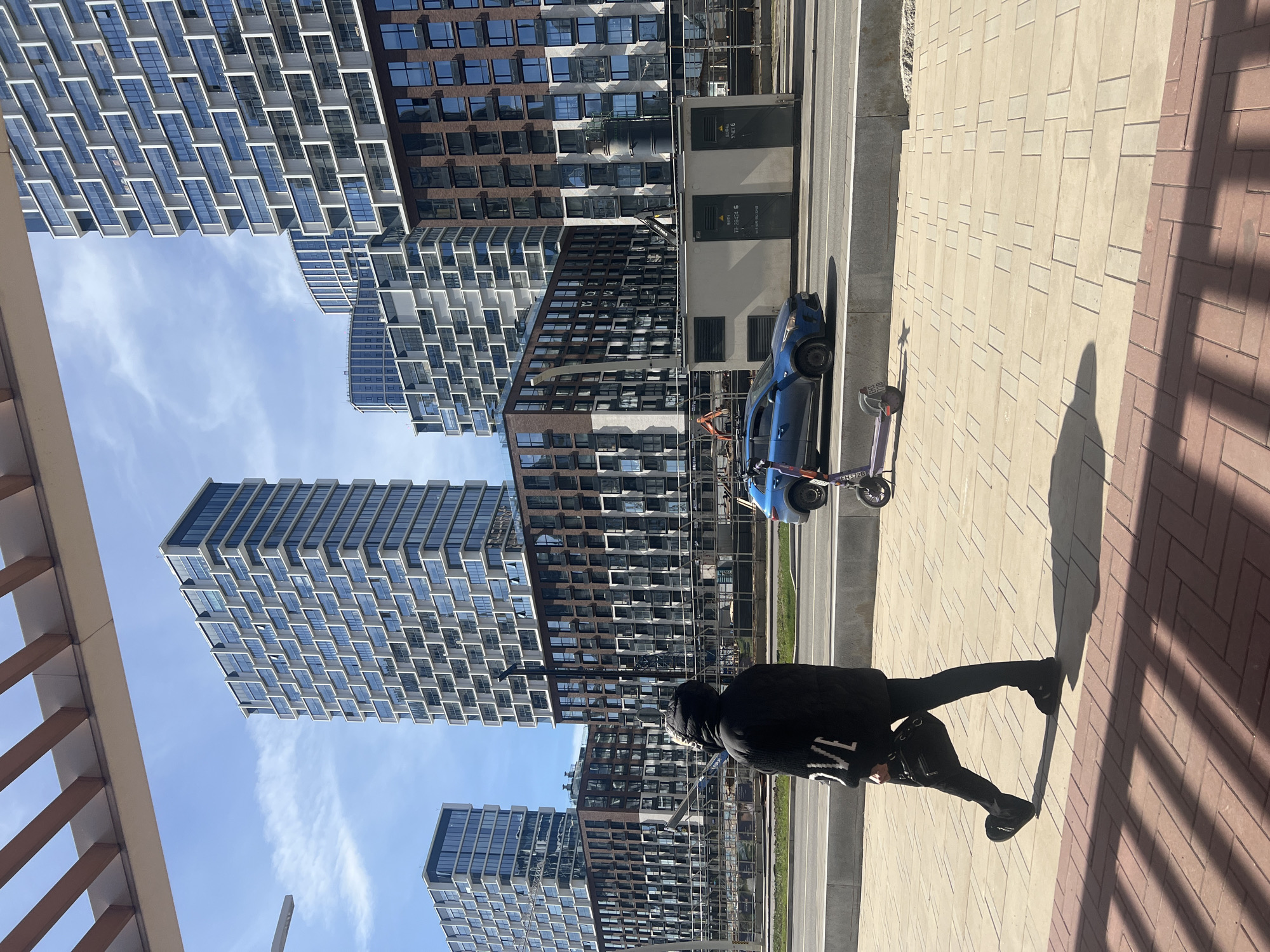 The image shows a street scene in Khimki, Russia. The background consists of several modern, high-rise apartment buildings under a clear, blue sky. Adjacent to the buildings is a road with a blue car parked along the side, and an electric scooter. A person wearing a black jacket is walking on the sidewalk. The sidewalk is made of light and red brick. Shadows are cast across the sidewalk.