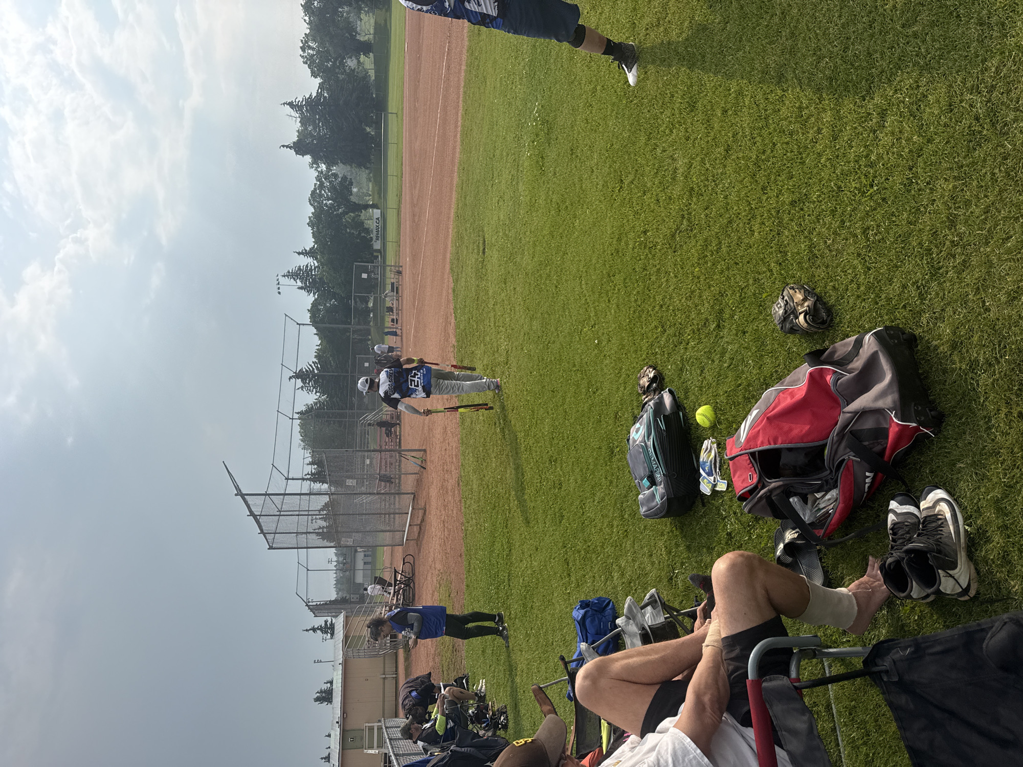 The image depicts a baseball or softball field on a cloudy day. In the foreground, several people are seated in chairs on the grass. One person, partially visible, is wearing a white t-shirt and black shorts, with athletic tape wrapped around their lower leg and ankle. Nearby are a pair of baseball cleats and several sports bags. Further back, a person in a blue jersey with the number 25 on the back is walking across the field, holding two bats. Other individuals are scattered around the field, and a fenced dugout area is visible in the background. The field itself has a dirt infield and grassy outfield. The overall setting suggests an organized sporting event. While the specific location in Calgary, Canada is not identifiable from the visual information alone, the presence of a baseball field with dugouts and spectators is consistent with a community sports park.