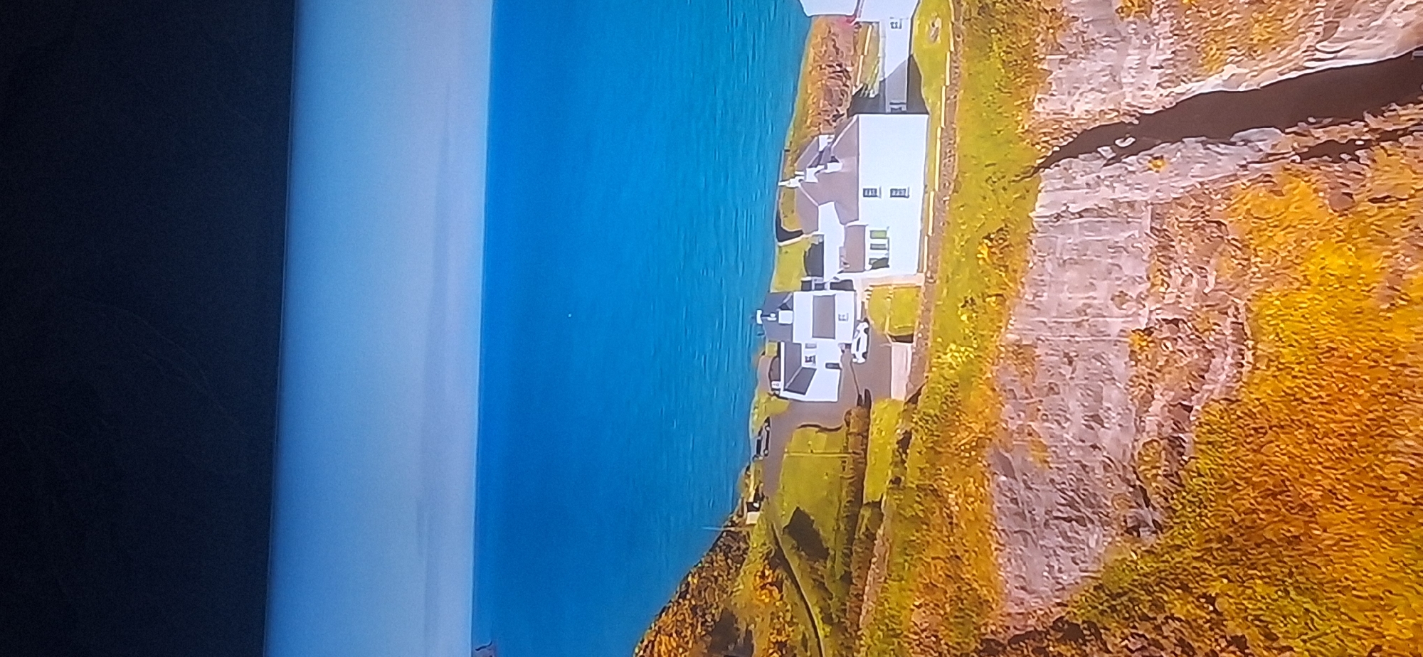 The image shows an aerial view of a coastal landscape. In the foreground, a dramatic cliff face is visible, covered in rough, textured vegetation that appears to be golden or yellow in color. A deep, dark crevice runs down the cliff.
Above the cliff, a green, grassy plateau stretches out towards the sea. Several white buildings with dark roofs are clustered on this plateau. The buildings appear to be houses or a small complex. A paved road runs past some of the buildings, and at least two vehicles, one white and one dark, are parked near the road. One of the buildings has a distinctive tower-like structure.
Beyond the plateau, a vast expanse of deep blue ocean meets a pale blue sky. The horizon is clearly defined. The overall impression is one of a remote, scenic location, likely in a temperate climate given the vegetation and clear skies. The image does not contain any specific landmarks or elements that would allow for identification of a location within the city of Bila Tserkva, Ukraine, as the environment depicted is characteristic of a coastal region, not an inland city.