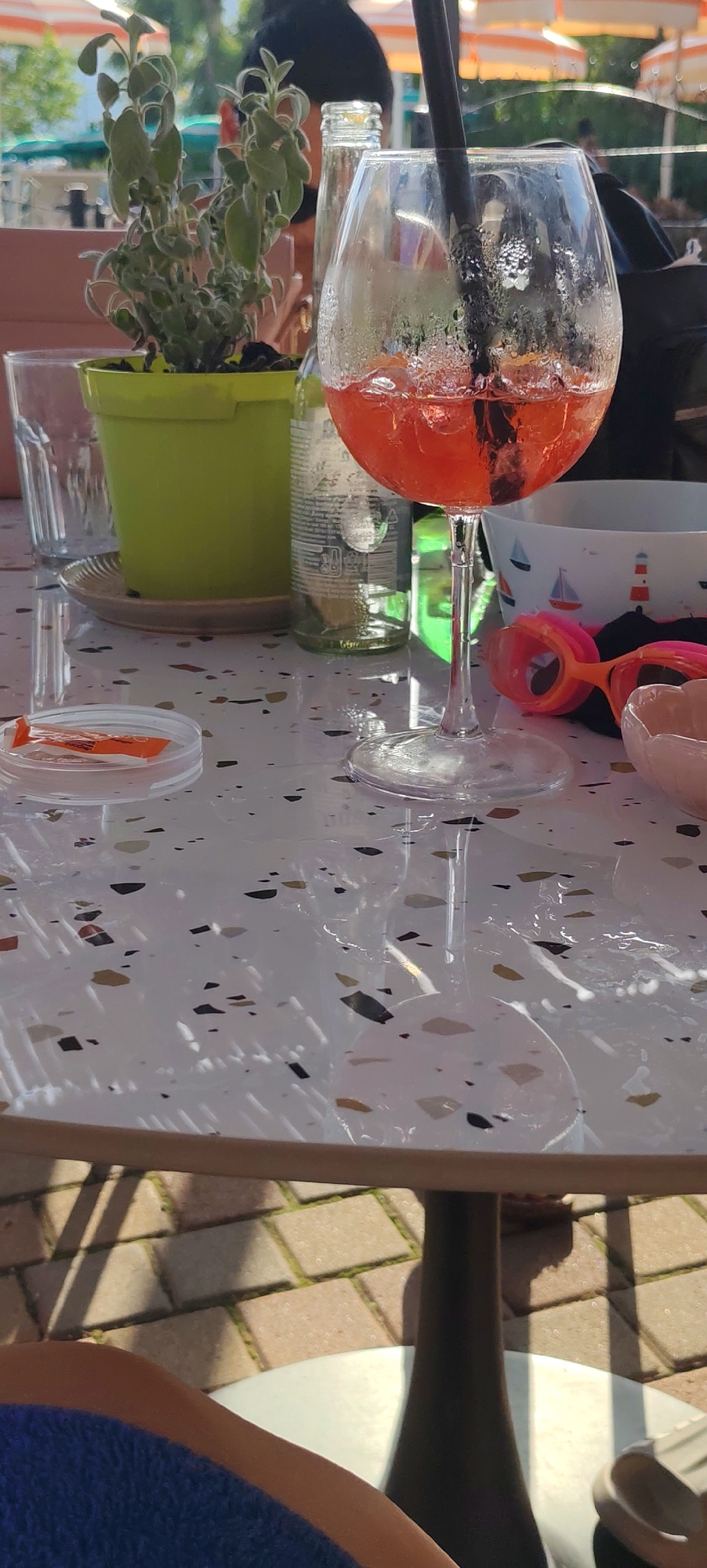 In the city of Luni, Italy, a tabletop scene unfolds, likely at an outdoor cafe or restaurant. A condensation-covered glass of red cocktail, adorned with a black straw, sits prominently, hinting at a refreshing drink on a warm day. Beside it, a clear glass and a green potted plant suggest a relaxed setting. Further elements like a glass bottle, a small dish containing orange items, and a patterned bowl with nautical motifs add to the everyday charm. Two pairs of brightly colored goggles, pink and orange, are casually placed, indicating a recent or impending visit to a swimming area, possibly a pool or the nearby sea. The table itself features a speckled terrazzo-like surface, reflecting the dappled sunlight that casts shadows from an unseen overhead structure. The background reveals hints of umbrellas and greenery, reinforcing the outdoor ambiance.