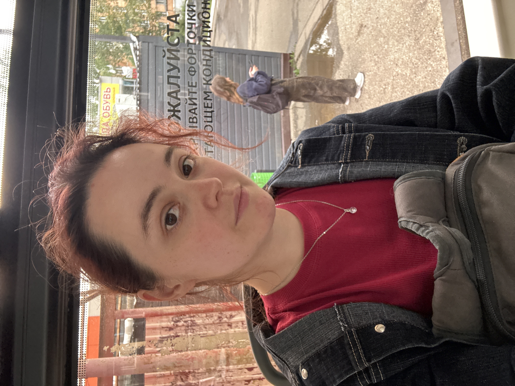 A young woman with reddish-brown hair is taking a selfie inside a vehicle, possibly a bus, based on the window view. She is wearing a red ribbed t-shirt, a dark denim jacket, and a silver necklace with a pendant. Her hair is somewhat messy and falls around her face. In the background, through the window, another person with brown hair is visible, wearing a dark blue sweater and brown pants, and carrying a backpack. They are looking at their phone. Outside the vehicle, there is a paved area with puddles, suggesting recent rain, and some buildings and trees are visible. A sign with Russian text, "ЖАЛУЙСТА" (Please), and another smaller sign with yellow lettering, "ОБУВЬ" (Shoes), are present on the glass. The location appears to be Izhevsk, Russia, given the context provided. The overall scene suggests a candid moment captured during travel or waiting.