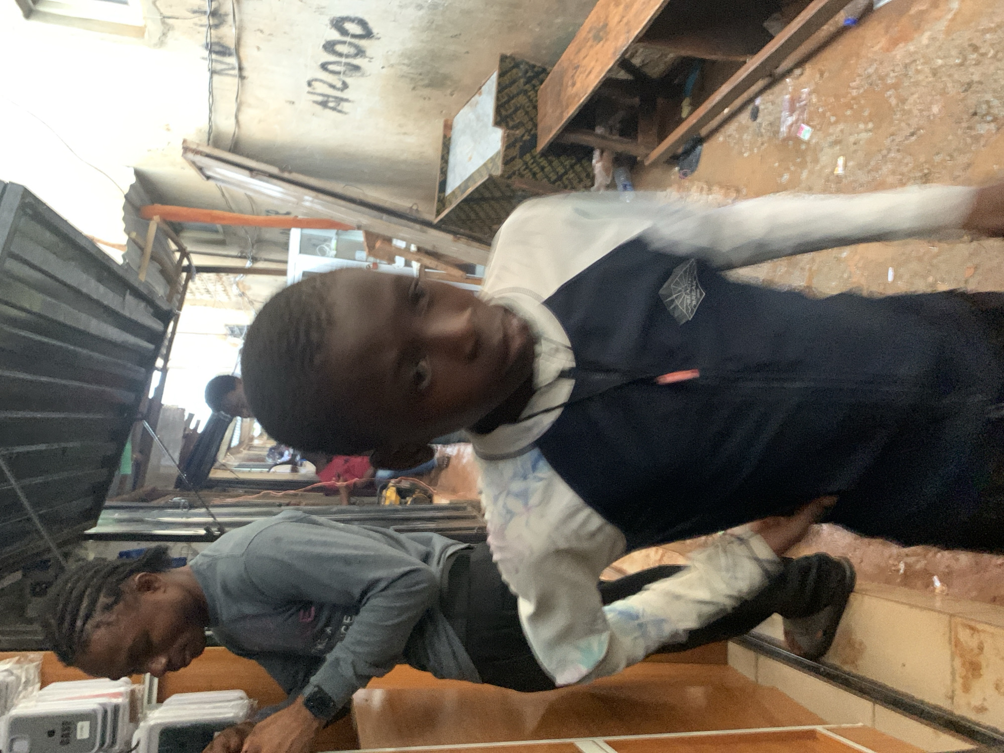 In Ado, Nigeria, a narrow outdoor market alleyway is captured. The ground is wet and muddy with scattered debris. On the left, a man with braided hair, wearing a grey long-sleeved shirt and dark pants, is attending to electrical meters mounted on a textured wall. To the right of the man, a young boy, the primary subject, is walking towards the camera, looking directly at it. He is wearing a blue and white long-sleeved shirt with a pattern on the sleeves. In the background, other people are visible, and stalls with various items and awnings suggest commercial activity. A concrete wall on the right side of the alley is marked with "N2000" in black paint. Wooden planks and shelves are visible, indicating the presence of goods or construction materials. The overall scene depicts a busy, informal marketplace environment.