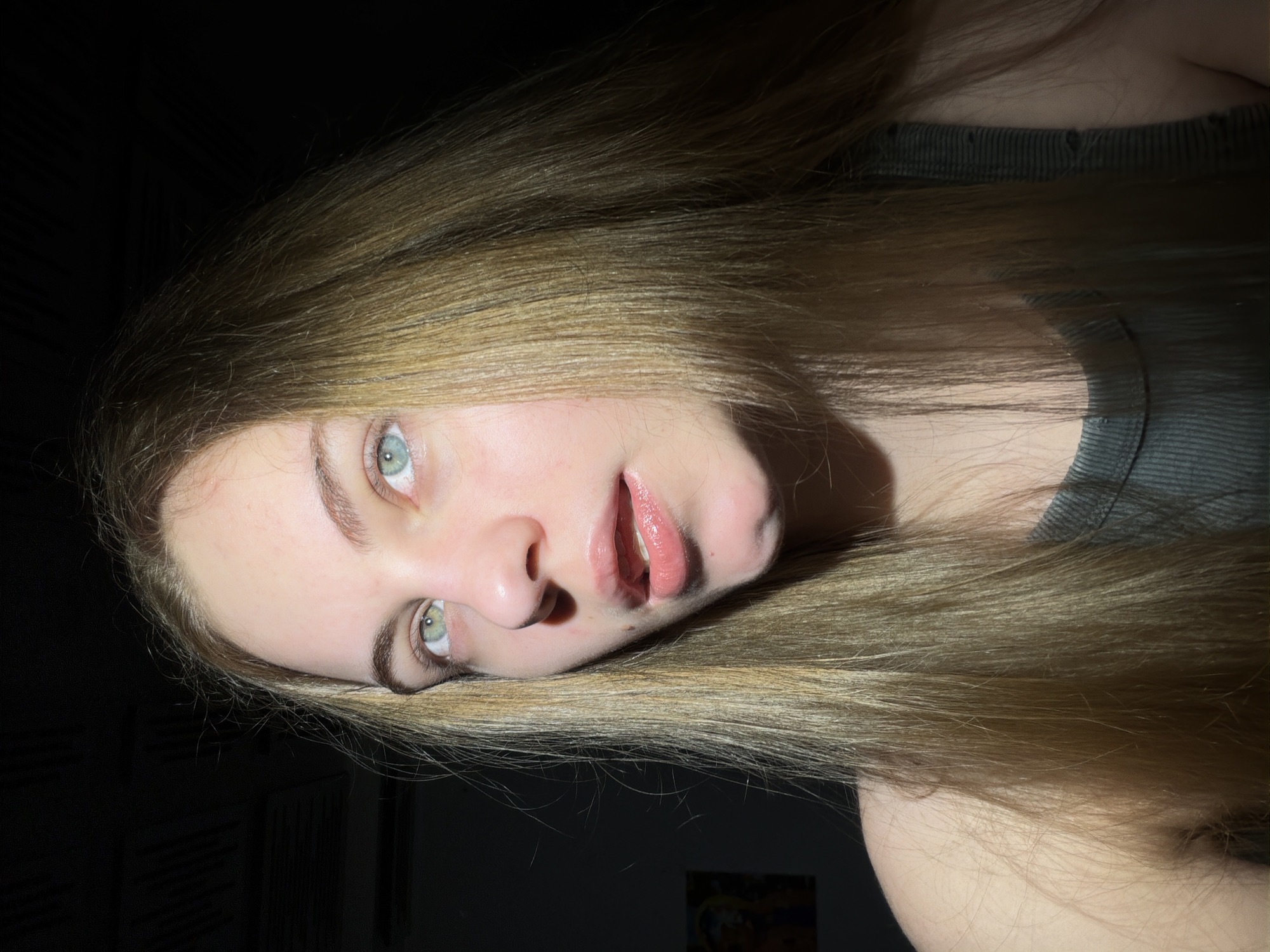 The image is a close-up, low-angle portrait of a young woman with long, blonde hair. She is looking upwards and slightly to her right, with her mouth slightly open. Her eyes are a striking blue-green. The lighting is dramatic, with a strong light source coming from the left, casting shadows across her face and hair. The background is dark, with some indistinct elements suggesting an indoor setting. A small portion of a framed artwork is visible on the left side of the background. The woman is wearing a dark, ribbed tank top. The image does not contain any information about the location within the city of Dnipro, Ukraine, nor does it depict any specific actions or interactions.