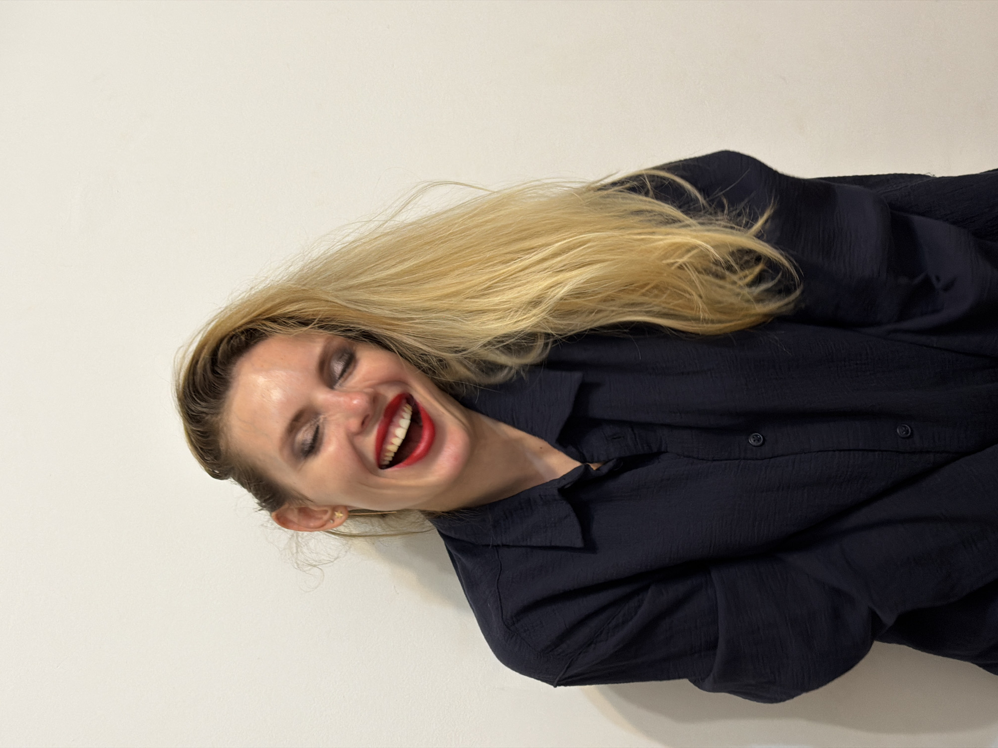 A woman with long blonde hair, wearing a dark button-down shirt, is laughing. She has red lipstick and her eyes are closed. The background is a plain, light-colored wall. There is no visible information indicating the location is Budva, Montenegro.