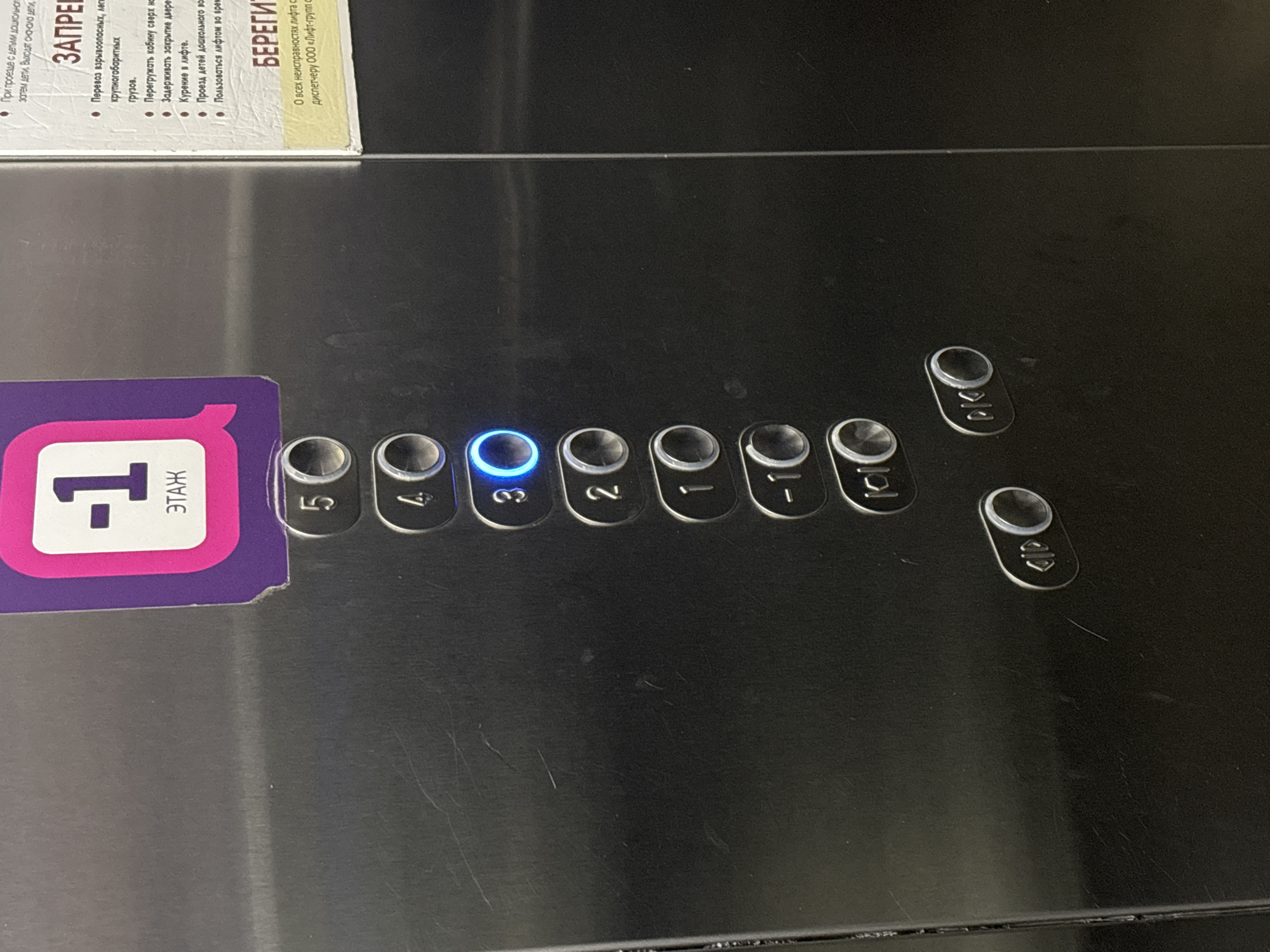 This image shows an elevator panel with floor buttons ranging from "-1" to "5," with the "3" button illuminated in blue. The panel also has buttons for opening and closing the doors. A sticker indicating "-1 этаж" (floor -1) is attached to the left side of the panel. Above the panel, there is a sign with safety rules and instructions in Russian. The sign includes the word "ЗАПРЕЩЕНО" (Forbidden) and "БЕРЕГИТЕ" (Take care). It also mentions ООО "Лифт-групп C." This indicates that it is likely located in the city of Perm, Russia because this company is located in Perm, Russia.