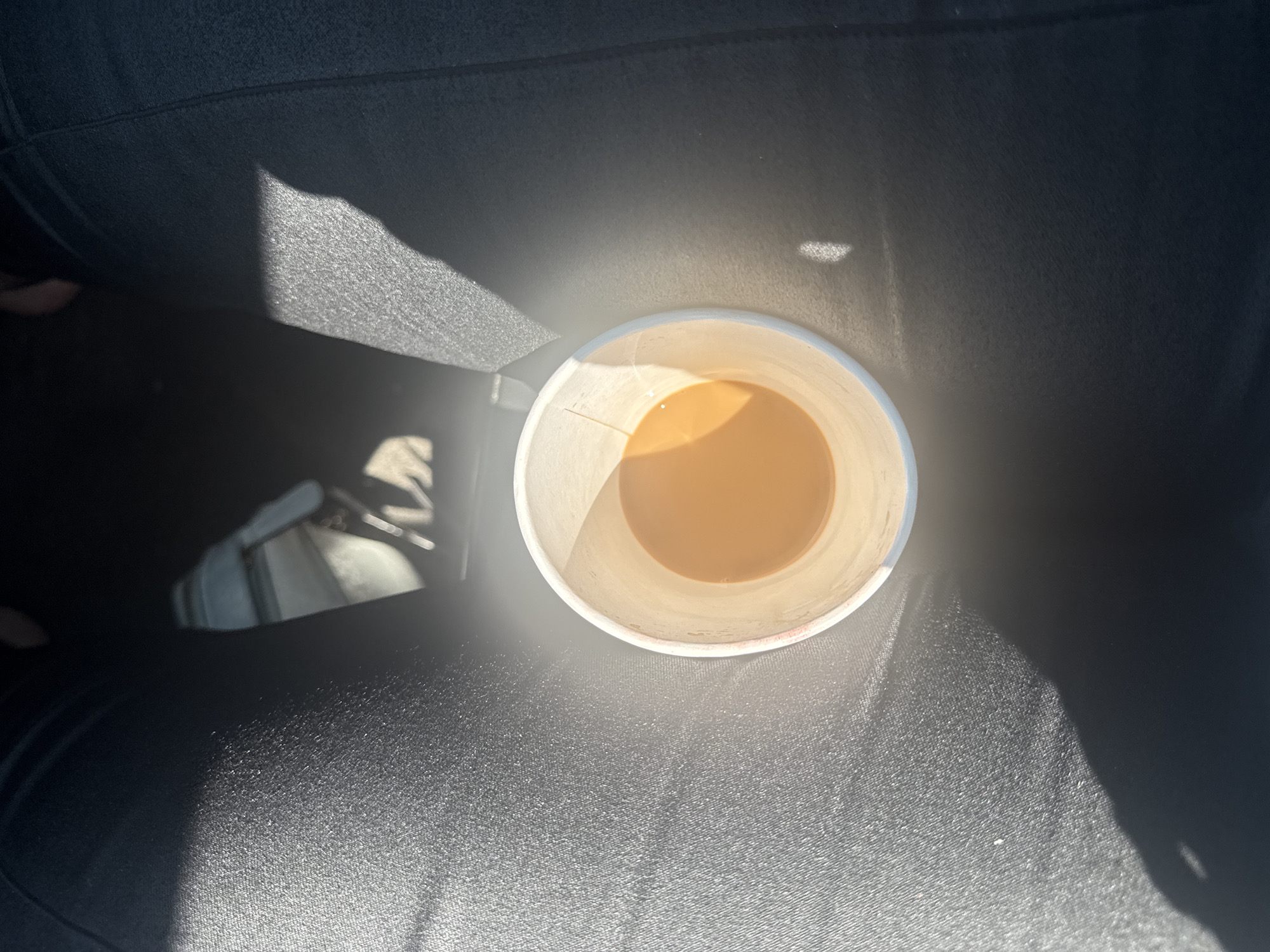 An overhead shot shows a white paper cup filled with a light brown liquid, likely coffee or tea, casting a shadow on a dark, textured surface. The cup is positioned slightly off-center to the right of the frame. The surface appears to be fabric, possibly leggings or pants, due to its texture and the way it conforms to the shape of the wearer's body. Sunlight streams in from the left, creating bright highlights and distinct shadows. In the background, out of focus, is a glimpse of what appears to be a car interior, with a white and grey seat. The image provides no information to suggest the location is the city of Calgary, Canada.