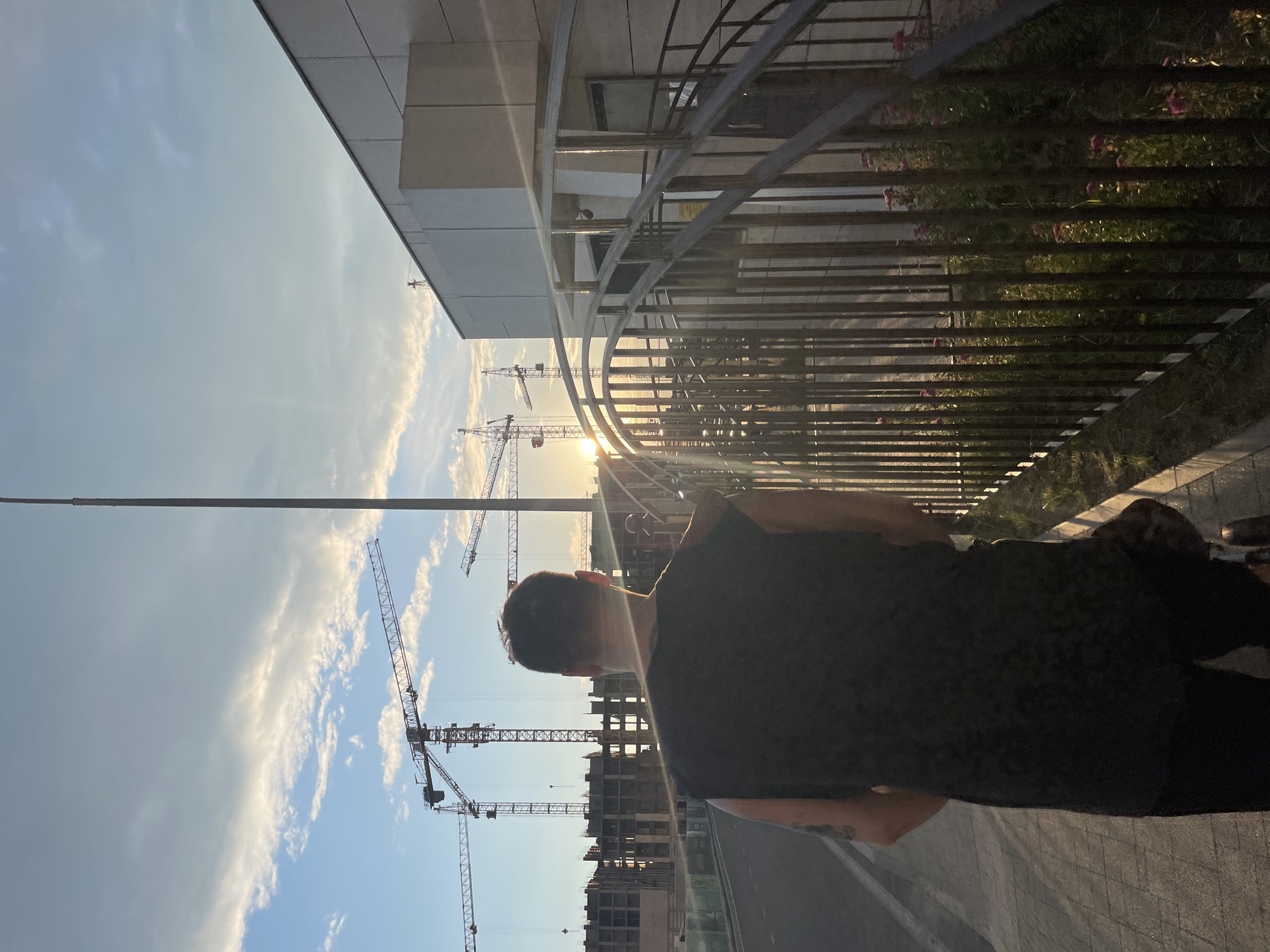 The image depicts a man walking away from the camera on a paved walkway. He is wearing a black sleeveless shirt and black pants. To his right, a metal railing separates the walkway from a patch of greenery with small pink flowers. In the background, several construction cranes are visible against a sky with scattered clouds, illuminated by the setting sun. A modern building with a light-colored facade is situated to the right of the frame. The scene appears to be a construction site in an urban or suburban area. The location is identified as Stavropol, Russia.