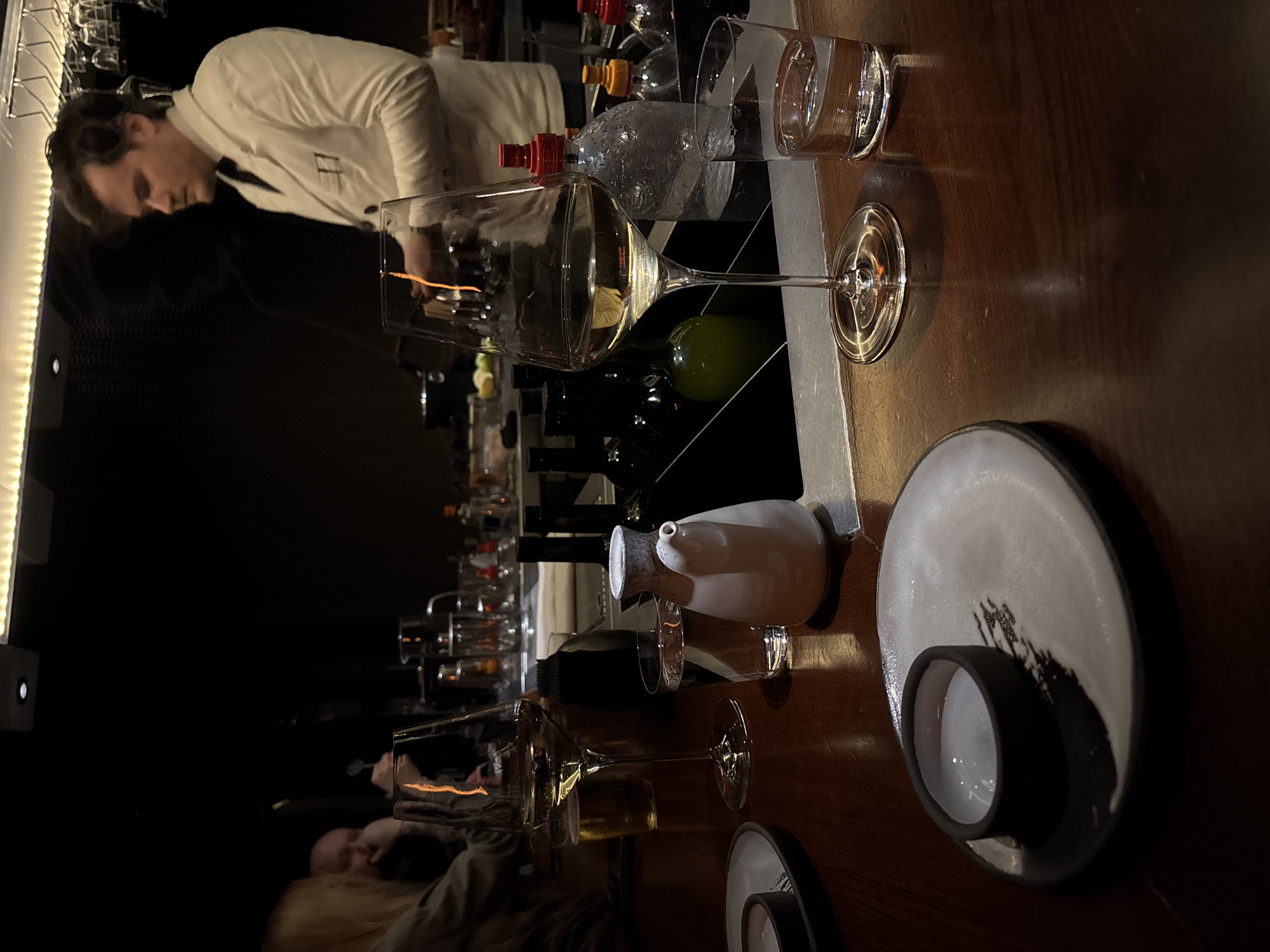 The image shows a dimly lit bar scene, likely in Moscow, Russia, featuring a bartender in a white jacket working behind a bar. On the bar, there are several wine glasses, one containing a clear liquid, likely white wine or water. A white porcelain sake bottle and a small sake cup resting on a white plate are also present. In the background, there are various bottles of alcohol and glassware on shelves. Patrons can be seen seated at the bar. The bar top is dark wood, and the overall atmosphere is sophisticated and intimate.
