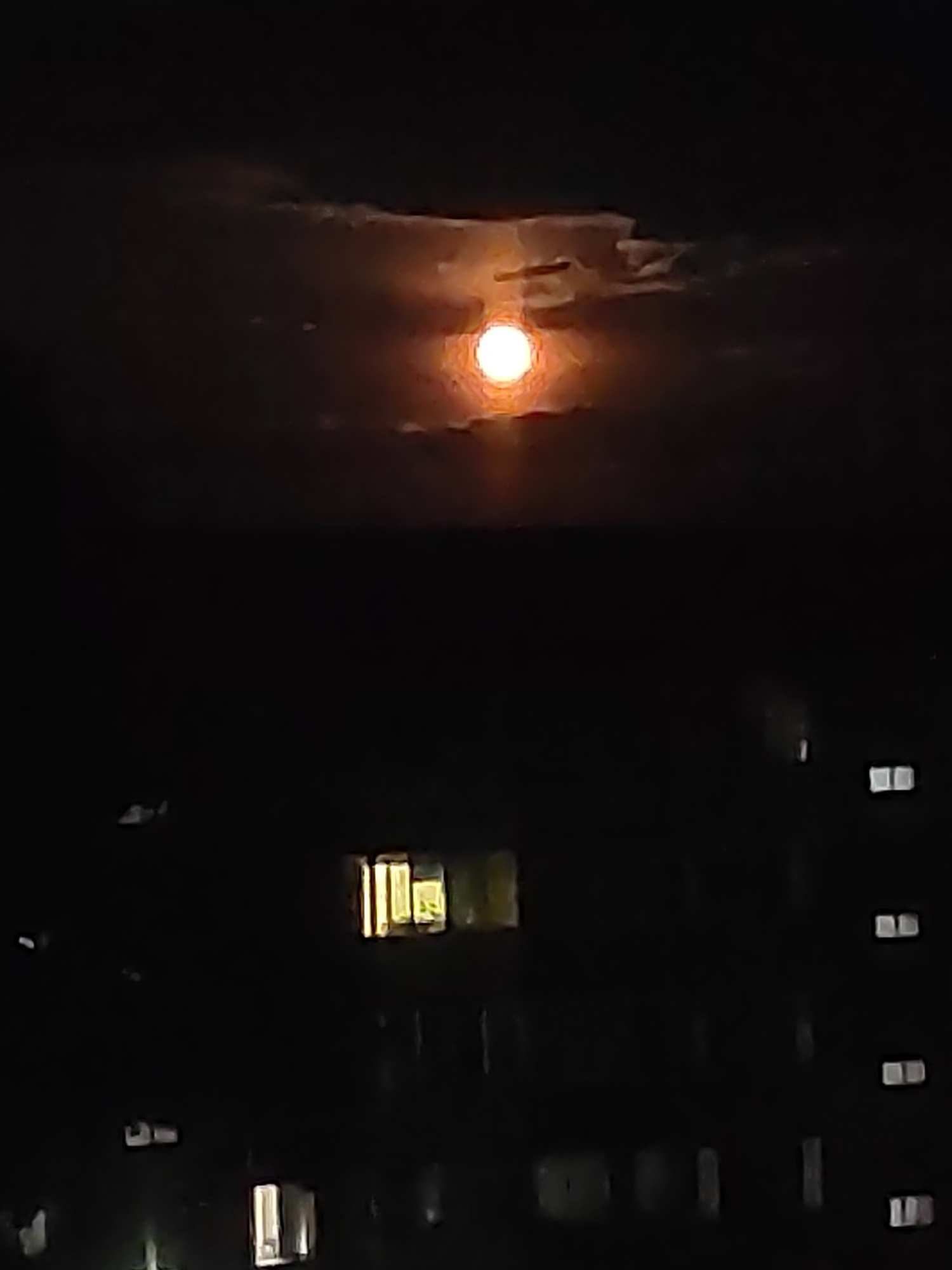 The image captures a night scene in Magdagachi, Russia. The primary subject is a bright, full moon peeking through dark, wispy clouds. The moon emits a warm, orange glow, casting a soft light. Below the moon, a silhouette of buildings is visible, illuminated by the moon and by lights from within some of the windows. The buildings appear to be residential or office blocks, with rectangular windows, some of which are lit with a yellowish hue, suggesting that people are inside. The overall atmosphere is tranquil and somewhat mysterious, evoking a sense of nighttime urban or suburban life under the glow of a celestial body. The time of day is clearly night, given the darkness and the presence of the moon and artificial lights. There are no people or animals explicitly visible, but the lit windows imply human presence within the buildings. No text is discernible in the image.