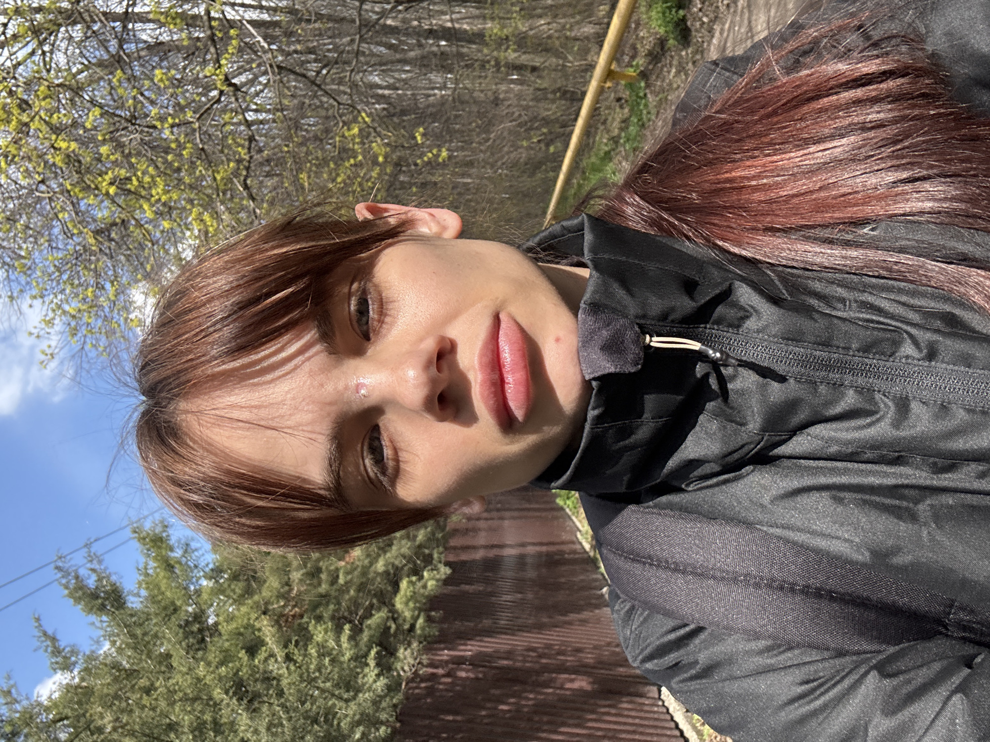 A young woman with brown hair is taking a selfie outdoors. She is wearing a black jacket. Behind her is a wooden fence, green trees, and branches with new spring leaves. The sky is visible in the upper left corner. The location cannot be determined with certainty to be Rostov-on-Don, Russia, as there is no specific evidence in the image to confirm this.