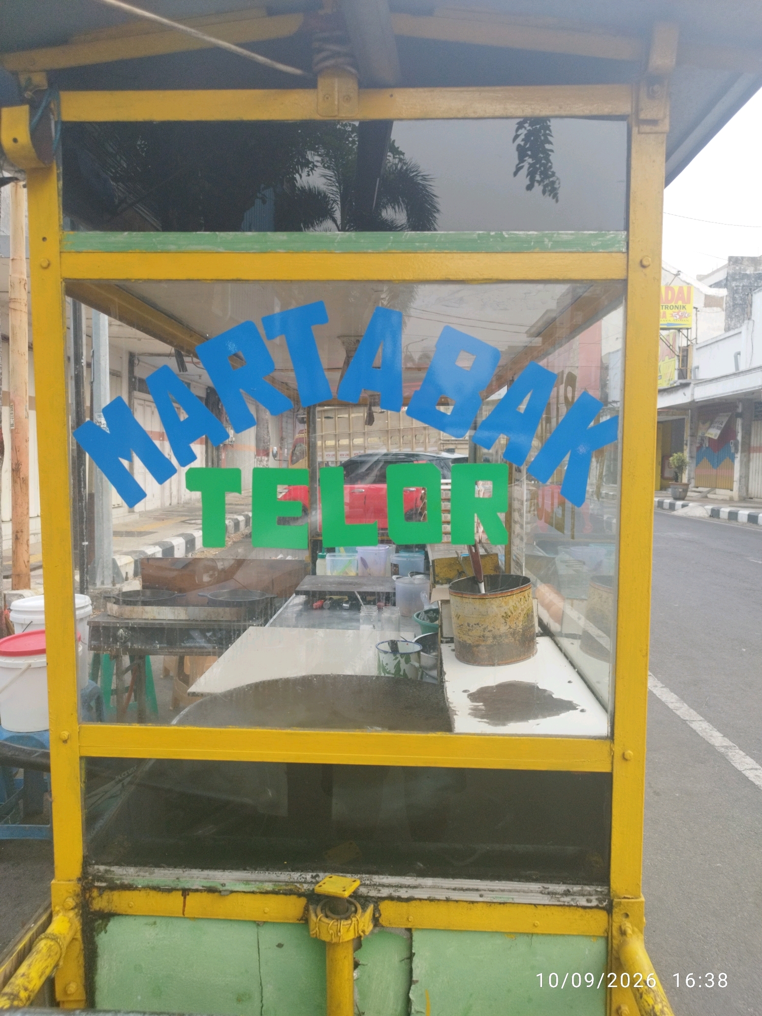 The image shows a food cart in Mojokerto, Indonesia. The cart has a yellow metal frame and green painted base. The front of the cart has a glass display with blue text reading "MARTABAK" and green text reading "TELOR." Inside the cart, various cooking utensils and containers are visible, including pans, buckets, and jars. The cart is parked on the side of a street with a sidewalk and buildings in the background. The timestamp "10/09/2026 16:38" is visible in the lower right corner.