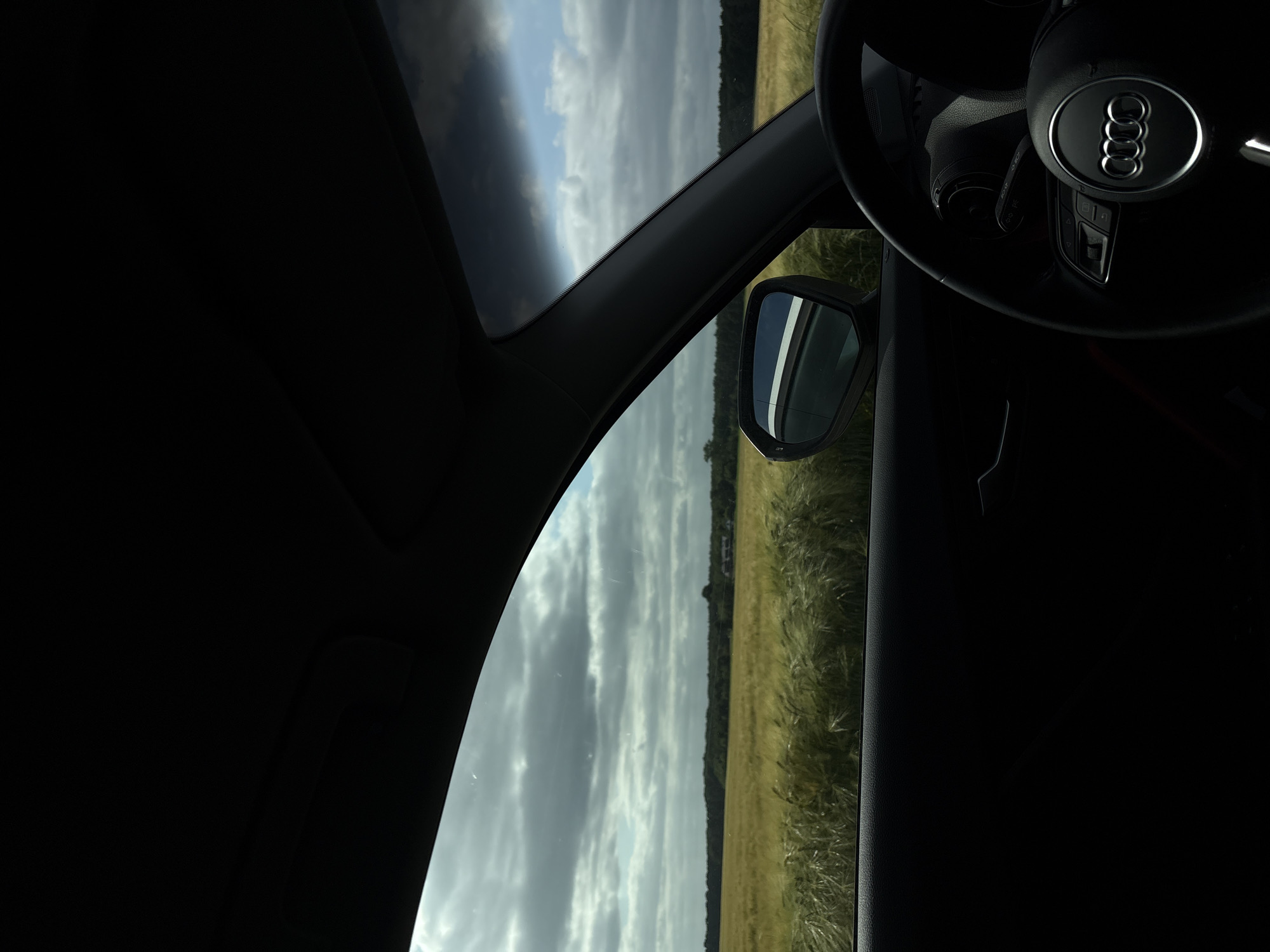 The image depicts a view from inside an Audi vehicle, specifically from the driver's seat, looking towards the left and front, in a rural area identified as Wegberg, Germany. The foreground shows the car's dark interior, including the top section of the dashboard, the driver's side door panel, and a portion of the black steering wheel with the silver Audi four-ring logo prominently centered. The car's side mirror is visible on the left, reflecting a white element and part of the sky.
Outside the car, a vast field of golden-yellow cereal crops, likely wheat or barley, occupies the mid-ground, extending horizontally across the frame. Beyond this field, a dark green line of dense trees or forest stretches across the horizon. Towards the right side, within or just beyond the tree line, a few reddish-brown structures resembling houses or farm buildings are faintly discernible. The sky above is partly cloudy, with large grey clouds dominating the upper left and central portions, while patches of blue sky are visible, particularly in the upper right. The scene is illuminated by natural daylight. No individuals are visible in the image.
