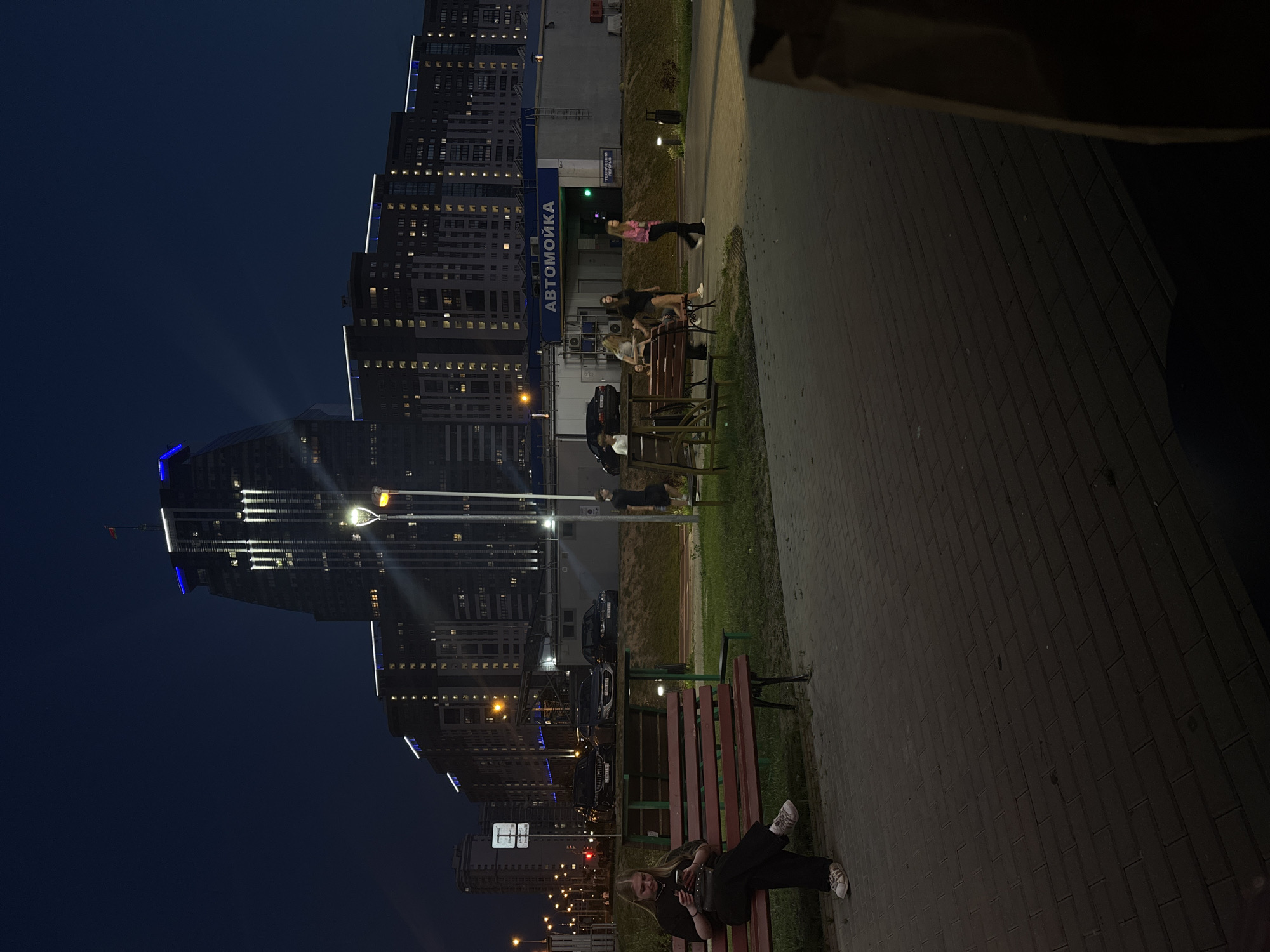 A woman sits on a park bench at dusk, looking at her phone. She is wearing black clothing and white sneakers. Behind her, a street with cars and buildings is visible. One of the buildings has "Автомойка" (Car Wash) written on it in large letters. The sky is dark blue, and lights from the buildings illuminate the scene. The overall setting suggests an urban environment in Minsk, Belarus.