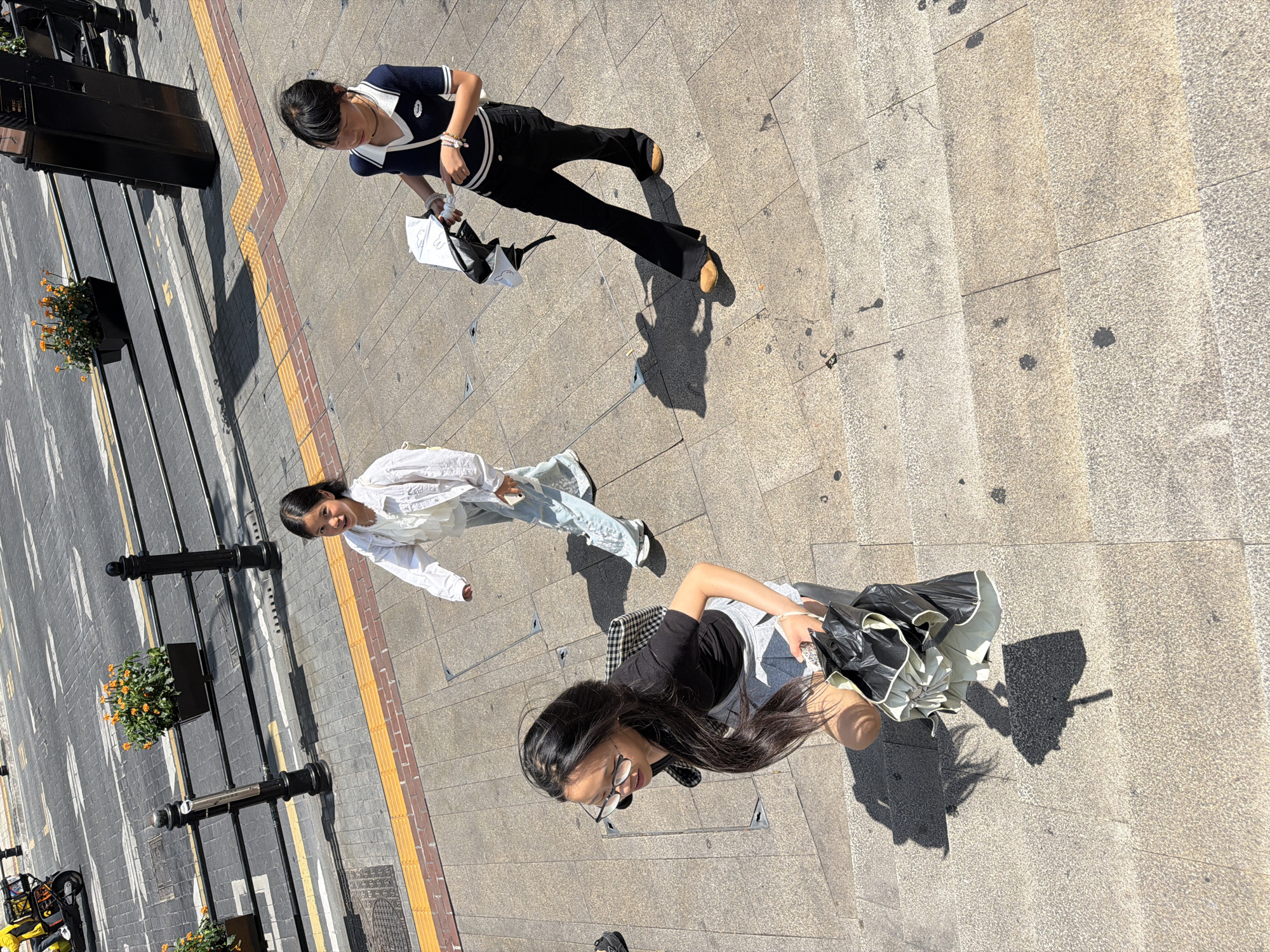 In Yuzhong District, China, on a sunny day, three young females walk up a stone staircase. The staircase appears to be outdoors in a public urban setting, with a street visible in the background. The street has trolley tracks and a yellow tactile paving strip.
The female in the foreground, on the left, wears glasses, a black t-shirt, and a grey skirt. She has long dark hair and is carrying a black umbrella and a checkered bag.
The second female, in the middle, wears a white jacket over a light-colored top and light blue pants. She is looking forward and appears to be walking.
The third female, on the right, wears a dark blue and white collared shirt, black flared pants, and light brown shoes. She is carrying a white and black umbrella.
The scene is captured from a high-angle perspective, looking down at the individuals. Shadows are cast on the staircase, indicating the direction of the sun. Orange flowers in black planters are visible along a railing in the background, alongside a street. A yellow scooter is parked on the street.