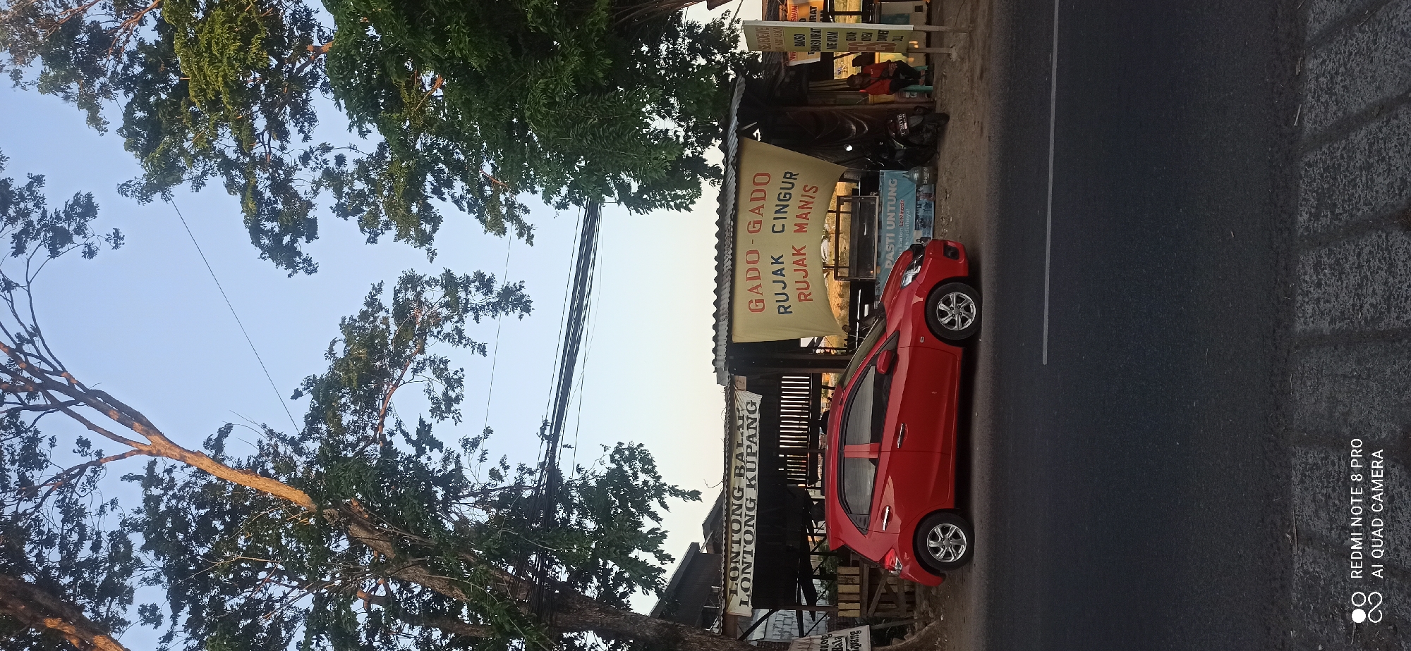 In Sedati Gede, Indonesia, a red Honda Brio car is parked on the side of a road, with a street food stall in the background. The stall has a banner displaying "GADO-GADO RUJAK CINGUR RUJAK MANIS" and "LONTONG BALAP LONTONG KUPANG", indicating the food items sold. A motorcycle is parked next to the stall. Two individuals are seated at the stall. Large trees with dense foliage flank the scene, and overhead power lines are visible. The sky is clear and light blue, suggesting it is daytime. The road in the foreground is asphalt, with a white dividing line. A drainage ditch with concrete curbing is present between the road and the stall. The overall scene depicts a typical roadside eatery in an Indonesian town.
