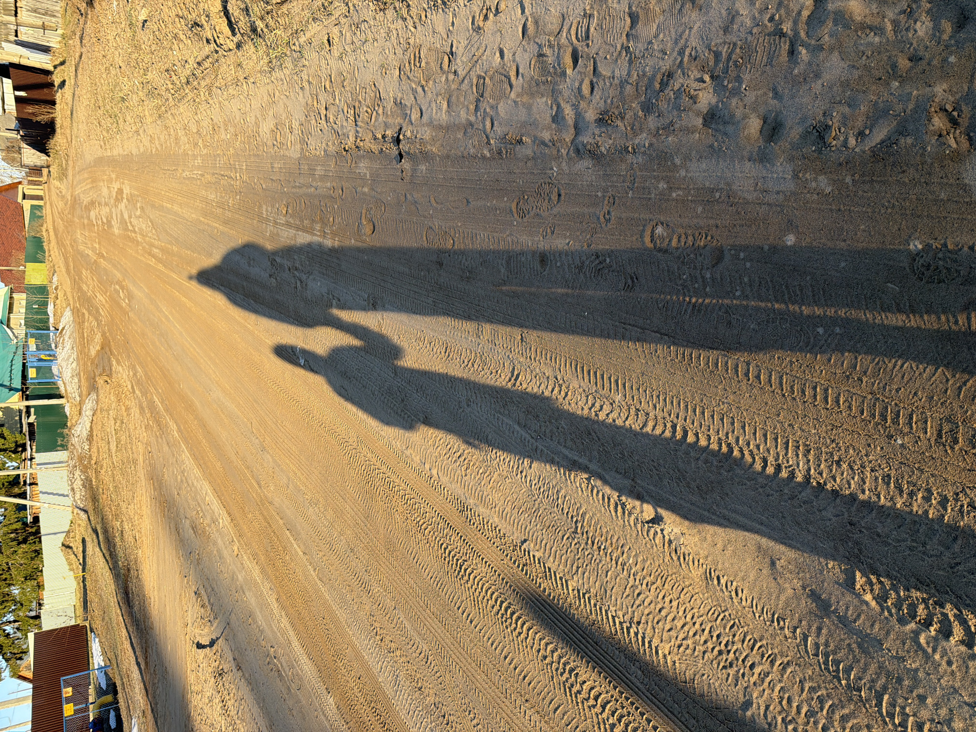 A high-angle, rotated, daytime view depicts a sandy road near buildings in Chaikovsky, Russia. The road surface displays multiple tire tracks and footprints. The long shadows of two people walking side-by-side are cast diagonally across the road. Buildings with varying roof colors are visible along the left edge of the frame. The environment appears arid with sparse vegetation.