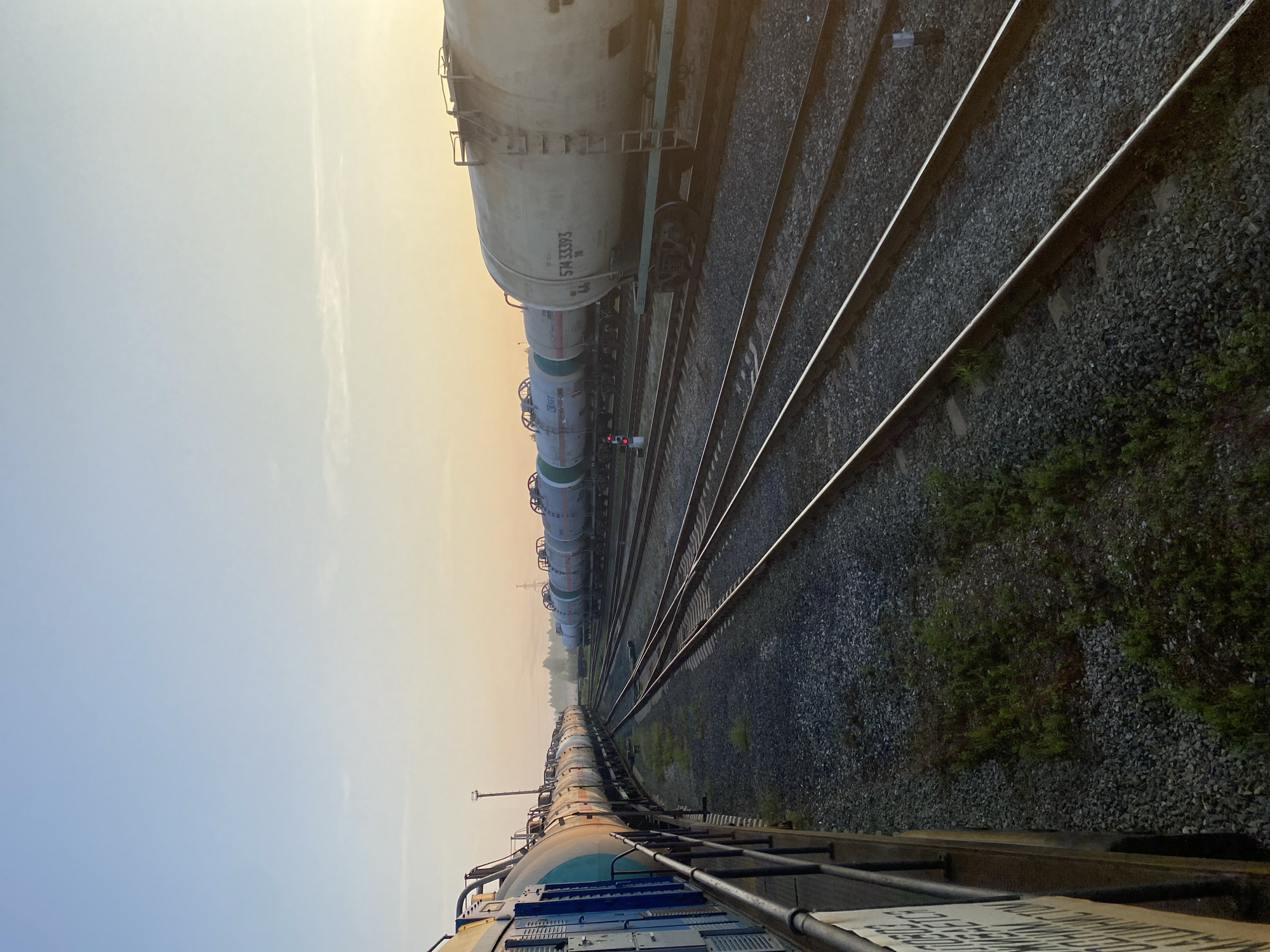 The image captures a train yard at Kauchuk Stantsiya, Russia, during what appears to be sunrise. Several tank cars are parked on multiple railway tracks that stretch into the distance. The sky is a clear gradient of light blue and soft orange, indicative of early morning light. On the left side of the frame, a section of a locomotive or train car with a blue and teal color scheme is visible, along with a railing and a sign with white lettering. The ground is covered with gravel and scattered green vegetation. In the background, trees and distant industrial structures are faintly discernible. There are no people visible in the image.