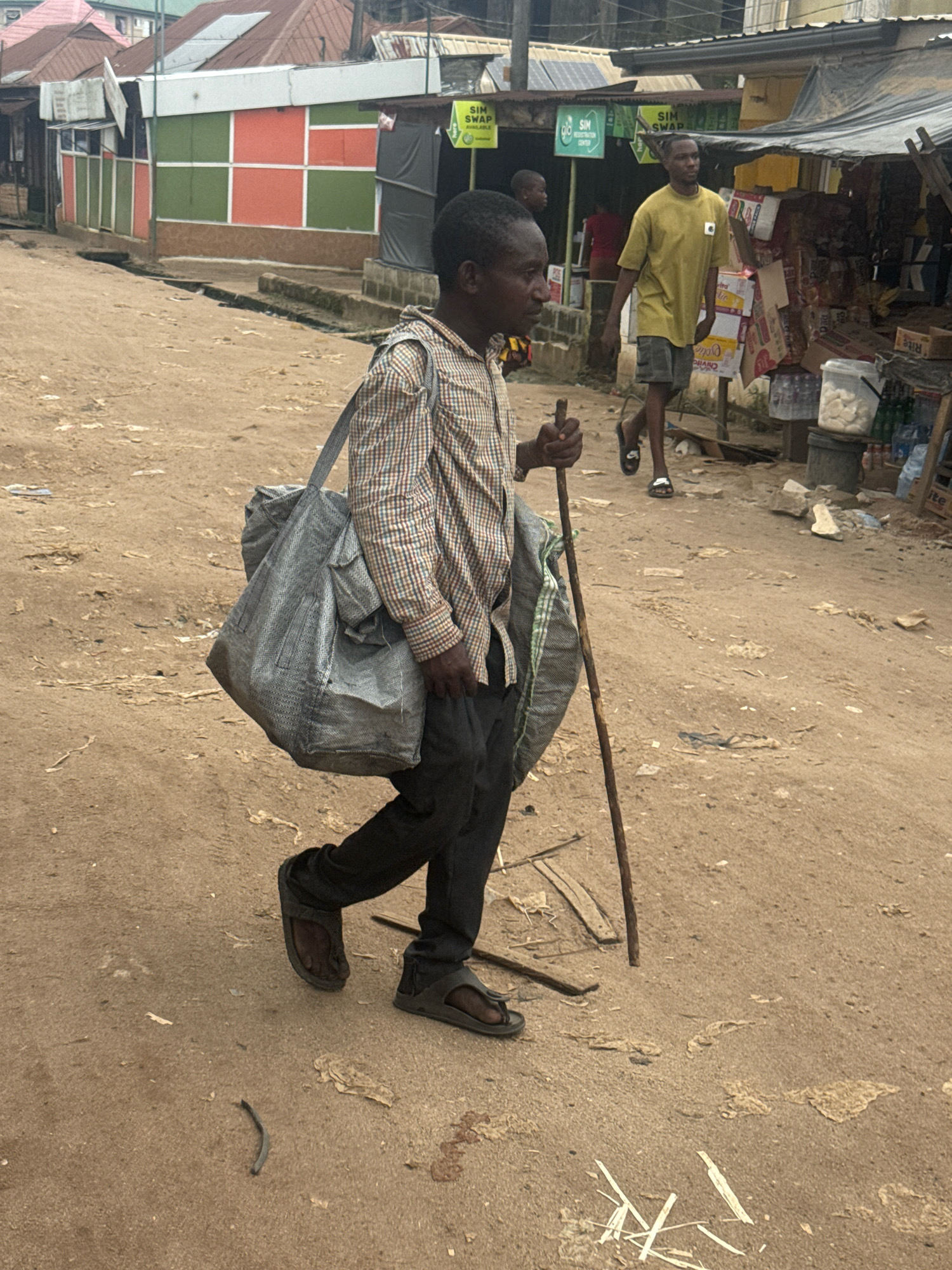 A man walks along a dirt road in what appears to be a market area. He is carrying a large, grey duffel bag slung over his shoulder and is using a walking stick. He is wearing a checkered long-sleeved shirt, dark pants, and sandals. In the background, another man in a yellow t-shirt and shorts is walking away from the camera. Shops with open fronts line the right side of the road, displaying various goods. Some of these shops have signs with text, including "SIM SWAP" and "SIM REGISTRATION CENTER". The buildings in the background have corrugated metal roofs and colorful facades. The ground is uneven and dusty. The lighting suggests it is daytime. The scene conveys a sense of daily commerce and movement in a bustling community.