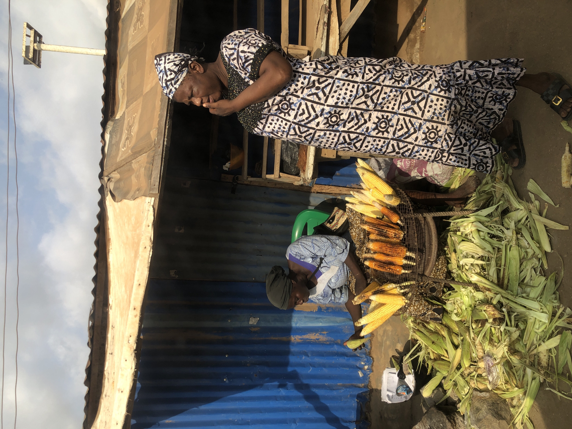 This image depicts a street scene in Otta, Nigeria, featuring two women selling corn. The woman in the foreground, wearing a black and white patterned dress and headscarf, stands facing right with her hand to her mouth, appearing to be in conversation. Behind her, a second woman in a blue and white patterned outfit and headscarf sits on a green chair, holding a piece of corn. She is surrounded by a large pile of corn husks and several ears of corn, some of which are roasted and displayed on a metal rack. The background shows a makeshift stall with a corrugated metal roof and blue siding. The environment appears to be an outdoor market, with the ground covered in dirt and debris. The sky above is partly cloudy.