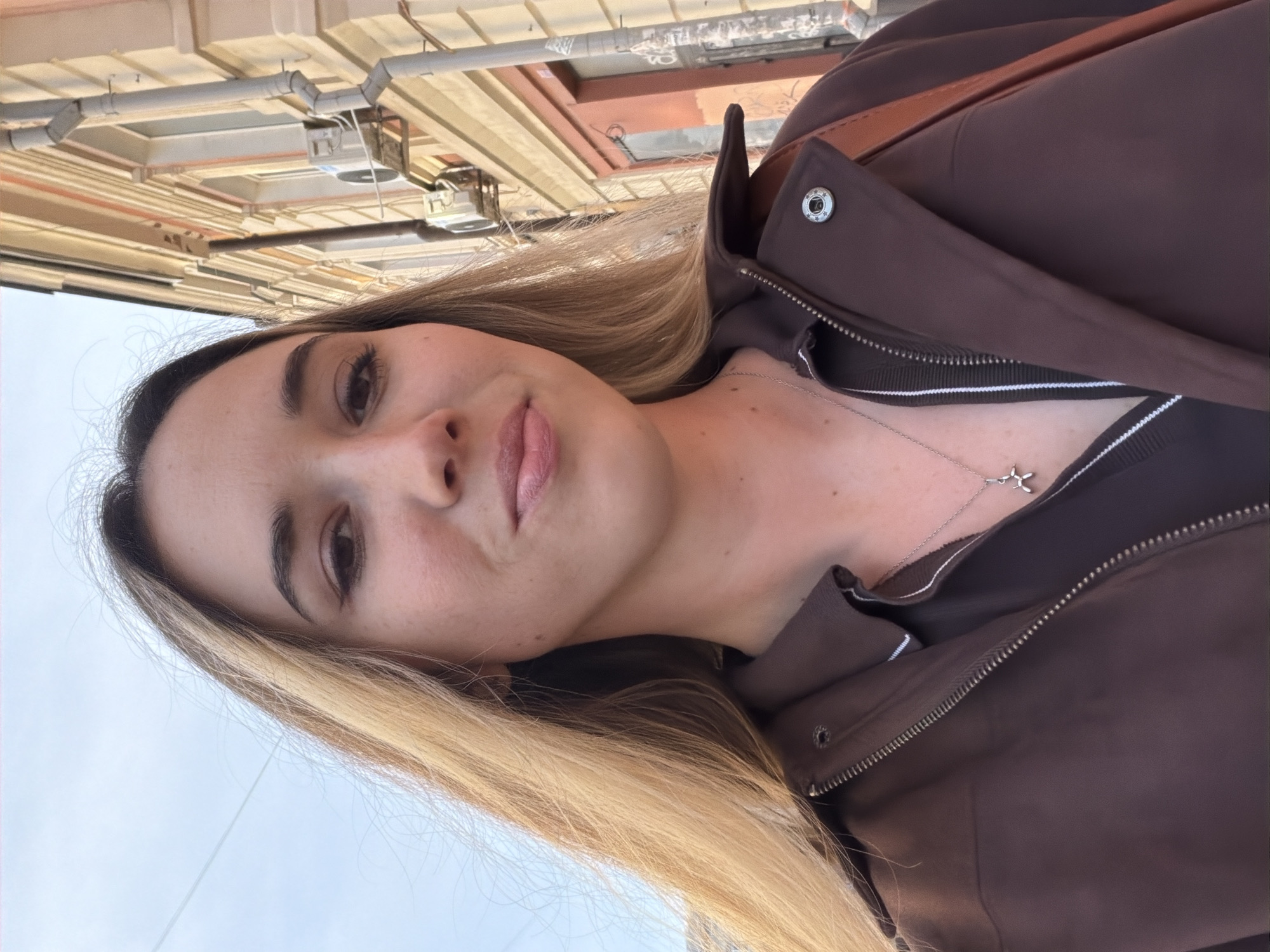 A young woman with long blonde hair and brown eyes is seen in a selfie taken outdoors. She is wearing a brown jacket and a silver necklace with a pendant. The background shows a light-colored building with architectural details and what appears to be pipes or utilities running along the top of the building. The perspective suggests the photo was taken from a slightly low angle. The exact location in Rostov-on-Don, Russia cannot be determined without further context.
