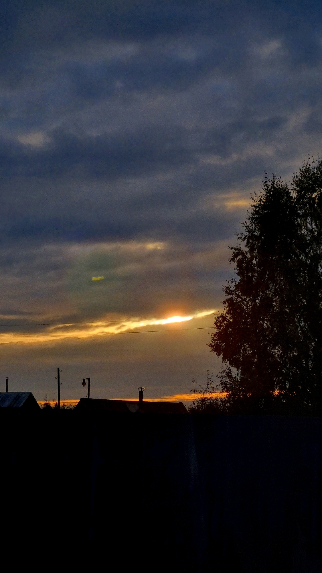 This is a vertical, medium-format photograph taken at dusk in the city of Vysokoe, Russia. The sky is filled with dark, heavy clouds, with a bright orange and yellow band of light cutting horizontally across the middle, indicating the setting sun. Silhouetted against this light are the dark shapes of several rooftops with chimneys, a utility pole with a lamp, and a large tree on the right side of the frame. In the foreground, a dark fence or wall runs horizontally, obscuring much of the lower half of the image. The overall mood is somber and quiet.
