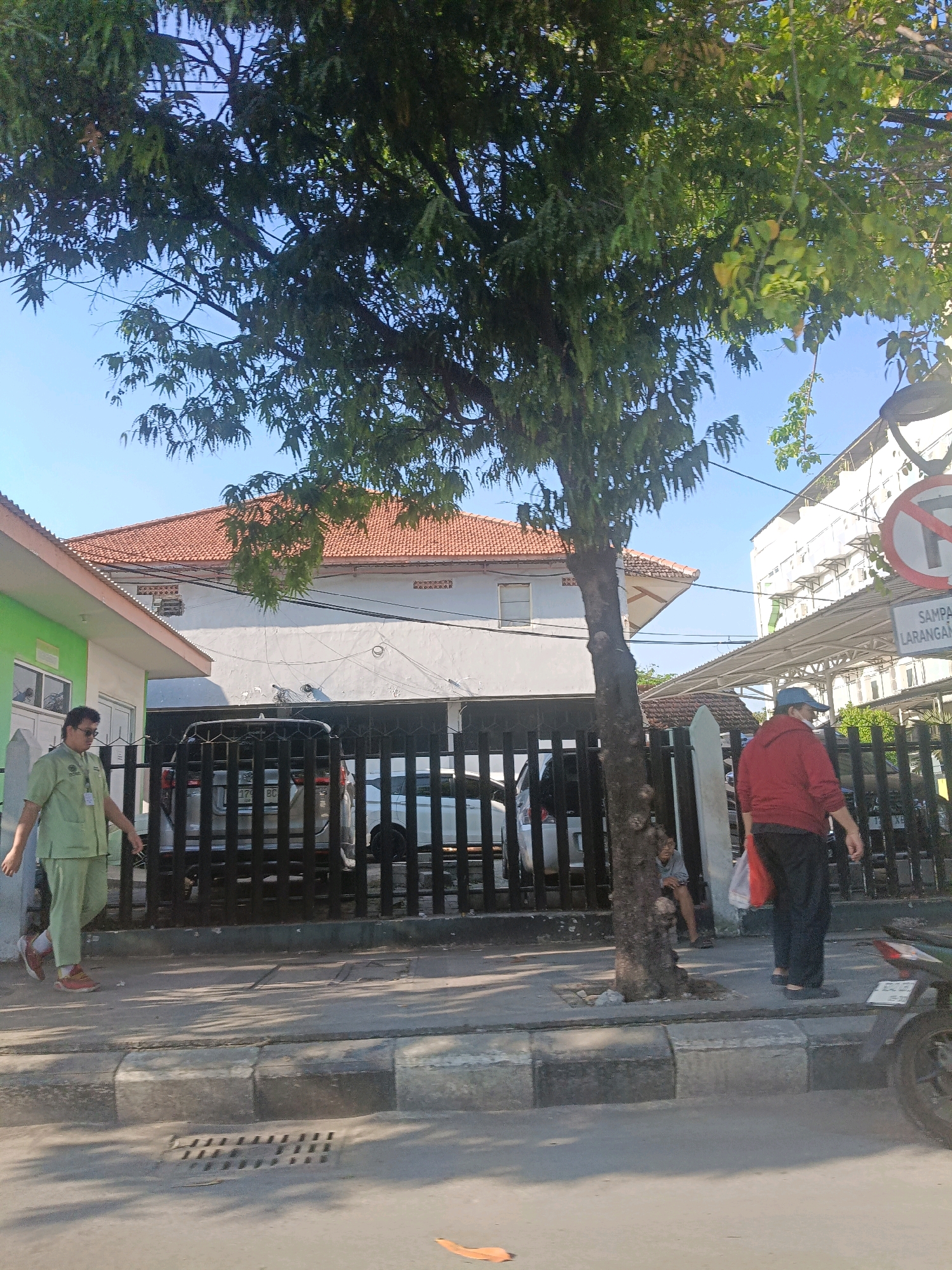 The image shows a street scene in Surabaya, Indonesia. On the left, a person in a light green uniform walks along a sidewalk. Behind them is a building with a green facade and a white tiled roof. A black metal fence separates the sidewalk from a property with a white building and a terracotta tiled roof. A silver SUV is visible behind the fence. Further to the right, another person in a red hoodie and black pants walks away from the camera, carrying a red bag. A street sign with a red circle and a diagonal line indicating "no entry" or prohibition is visible on a pole to the right, above a sign that reads "SAMPAI LARANGAN" which translates to "Until Prohibition." Trees with green leaves line the street. A motorcycle is parked on the right side of the image. The sky is clear and blue. The overall impression is of a bright, sunny day in an urban environment.