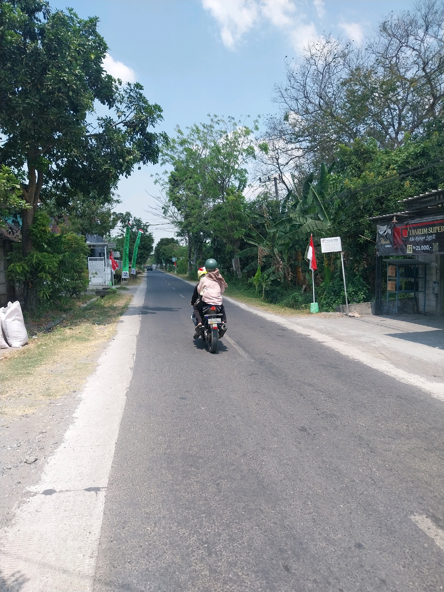 A two-person motorcycle travels down a paved road in the city of Campur, Indonesia. The driver, wearing a green helmet and a pink shawl, navigates the road, while the passenger, also wearing a pink shawl, sits behind them. The motorcycle has a blue license plate with white lettering. Trees line both sides of the road, with lush green foliage and some bare branches reaching towards a bright blue sky with scattered clouds. Several white sacks are piled on the left side of the road, and a building with a dark awning and a sign that reads "DIARUM SUPER" is visible on the right. A red and white flag, likely the Indonesian flag, stands in front of the building. The road itself is gray asphalt with a white line marking the center. The scene appears to be a typical day in a rural or suburban area of Indonesia.