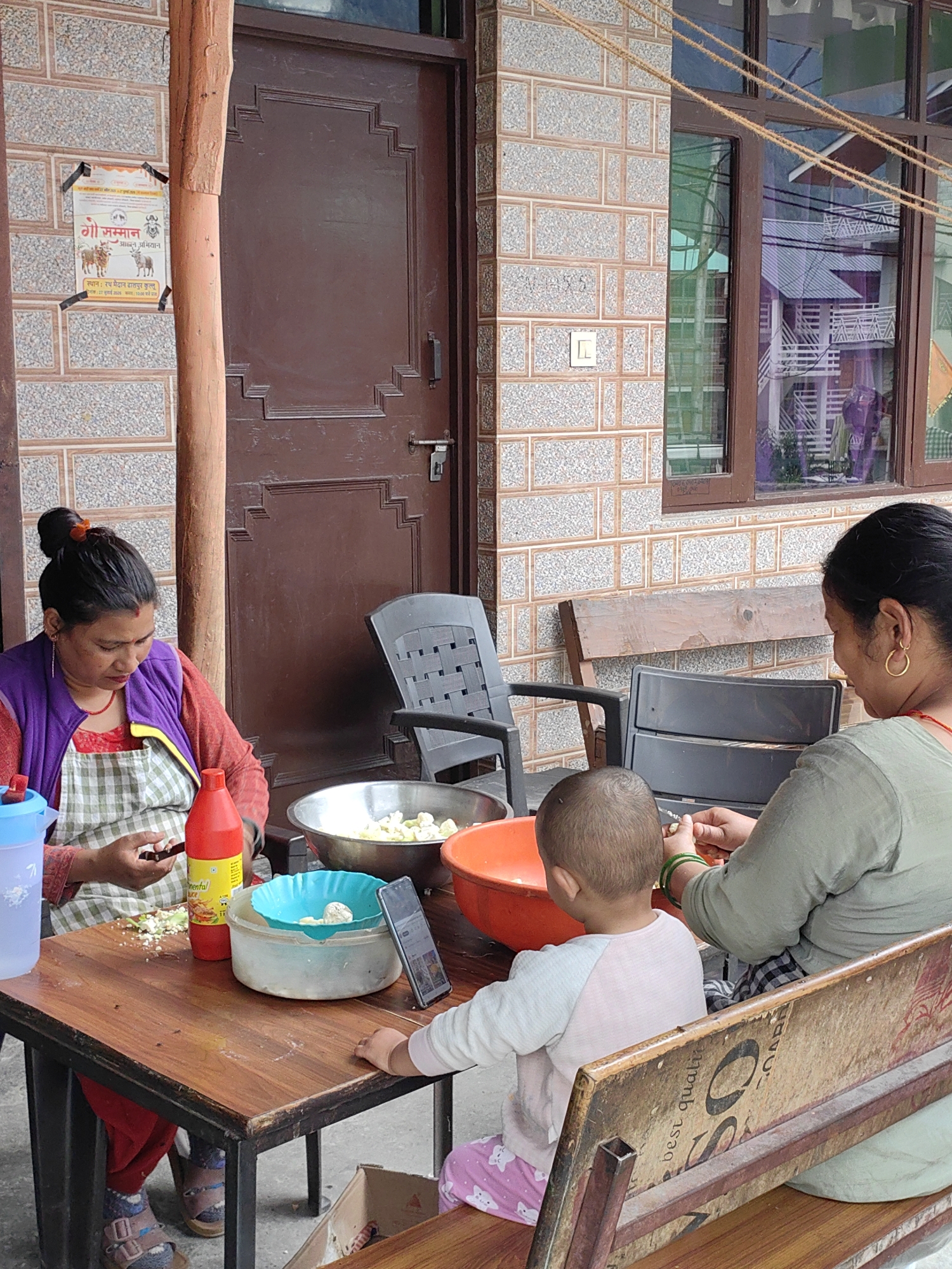 In Barheta, India, a group of individuals are gathered around a wooden table, engaged in food preparation. A woman, wearing a purple vest over a red shirt and a checkered apron, is seated on the left, focused on her mobile phone while chopping vegetables on the table. To her right, a red bottle of "Continental Sauce" stands upright. In front of her, a large metal bowl contains chopped cauliflower. A blue colander sits atop a white bowl, with cauliflower florets inside. A mobile phone is propped up next to the colander.
A young child, wearing a white and pink outfit, is seated in front of the table. To the right of the child, another woman, wearing a green shirt, is also engaged in food preparation, possibly peeling or cutting vegetables. A large orange bowl is situated behind the child, and a wooden bench is visible in the foreground, partly obscuring a cardboard box. The background features a building with a brown door and windows, constructed with brickwork and tiles. The overall scene suggests a casual, communal activity taking place outdoors.