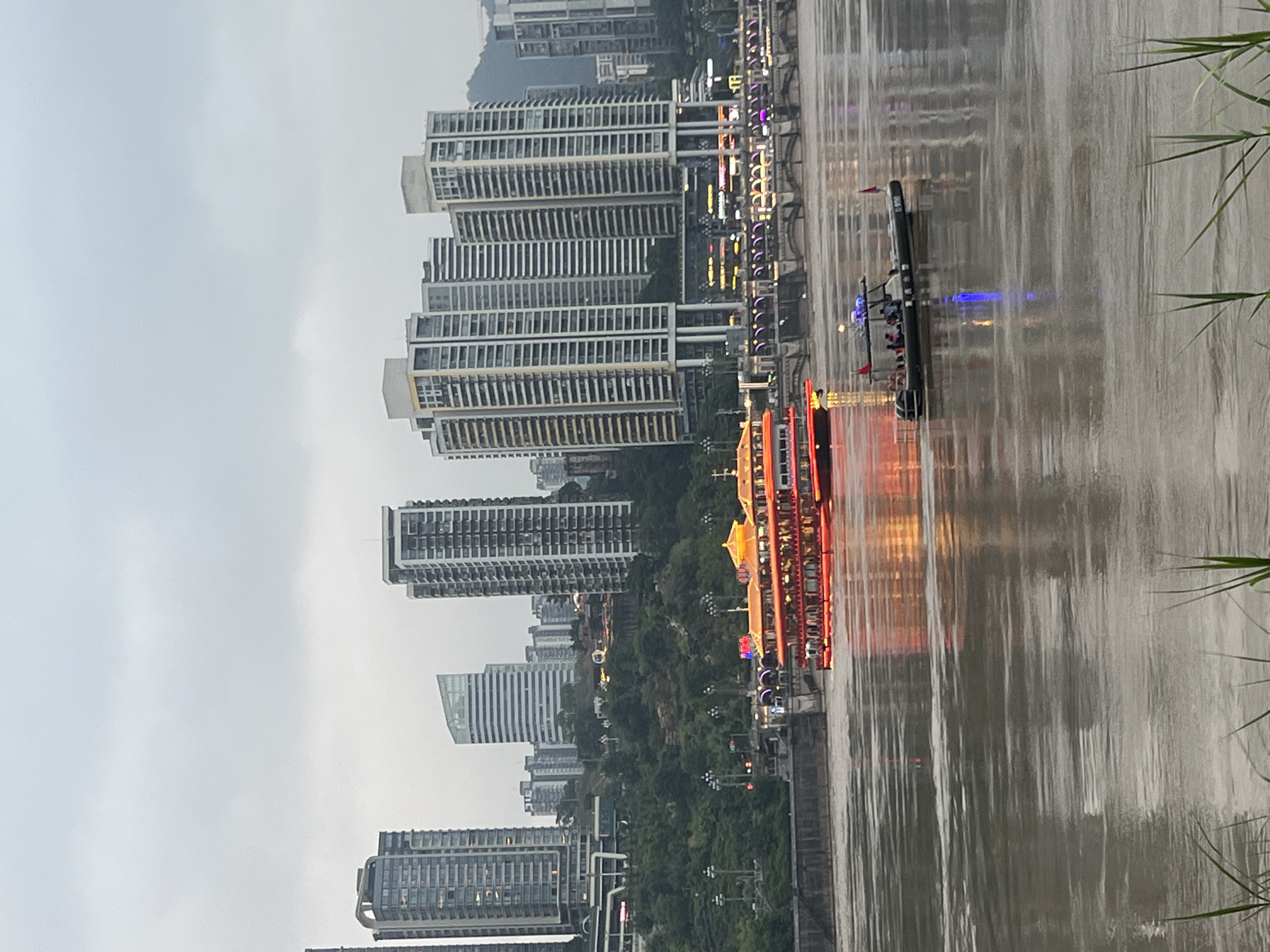 This photograph depicts a wide, murky river in the Nan'an District of Chongqing, China, under an overcast sky. In the foreground, a small, dark-colored boat with several occupants and a blue light on its roof navigates the water. A larger, multi-decked tour boat, illuminated with bright red and orange lights, is moored or traveling slowly on the river. The background features a densely built urban landscape with numerous tall, modern skyscrapers and a prominent bridge spanning the river. The riverbanks are lined with retaining walls, and green foliage is visible on the hills behind the buildings. The overall impression is one of a busy, illuminated city at dusk or dawn, reflected in the rippling surface of the river.
