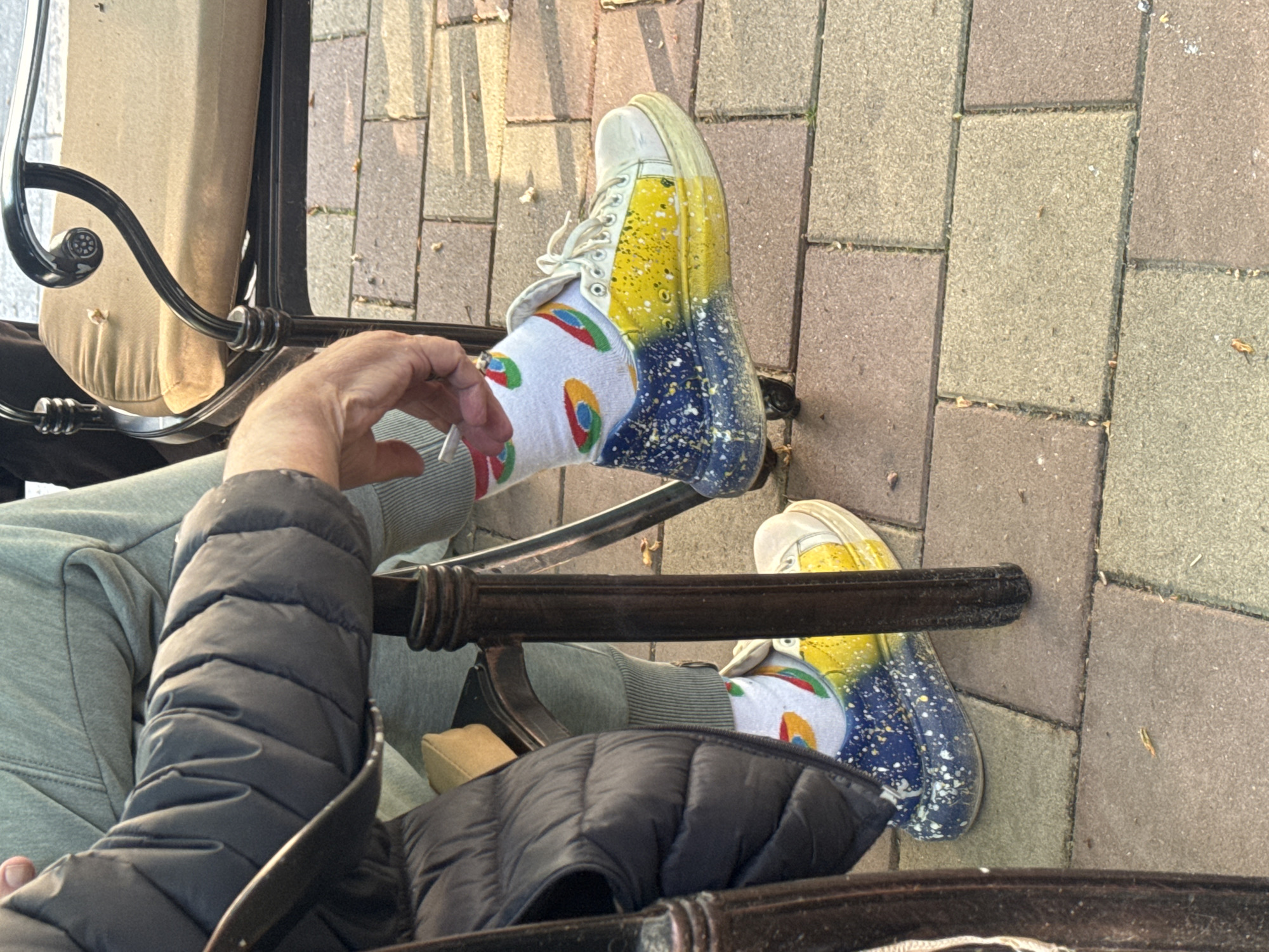 A person wearing yellow and blue splatter-painted sneakers and socks with colorful Google Chrome logos is sitting in a chair outside. The ground is paved with tan and brown rectangular stones. The person is wearing light gray sweatpants and a puffy black coat. The scene is outdoors in daylight. Location details are not deducible from the image.