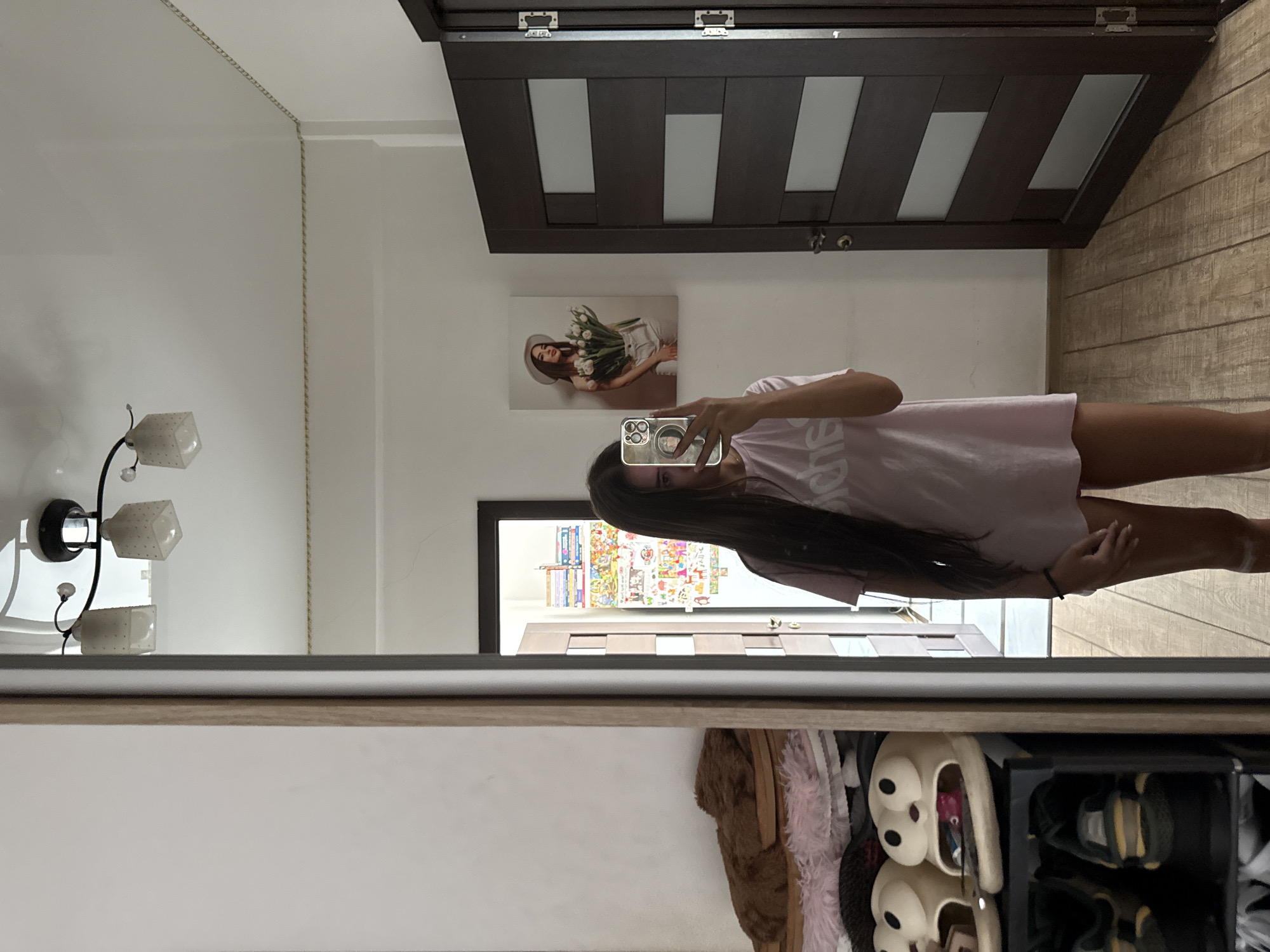 A young woman with long dark hair, wearing a pale pink t-shirt and shorts, stands in a room and takes a mirror selfie. She is holding a smartphone in her right hand and her left hand is resting on her thigh. The room appears to be a bedroom or dressing area. To her left, a large mirror reflects a shelf unit filled with various items, including fuzzy slippers and shoes. To her right, a dark wooden door stands ajar, revealing a glimpse of another room with what appears to be colorful posters on the wall. Above the woman, a light fixture with multiple glass shades is attached to the ceiling. A framed picture of a woman holding a bouquet of white tulips hangs on the wall to her right. The flooring is made of light-colored wooden planks. The scene is domestic and appears to be in a residential apartment in Chișinău, Moldova.