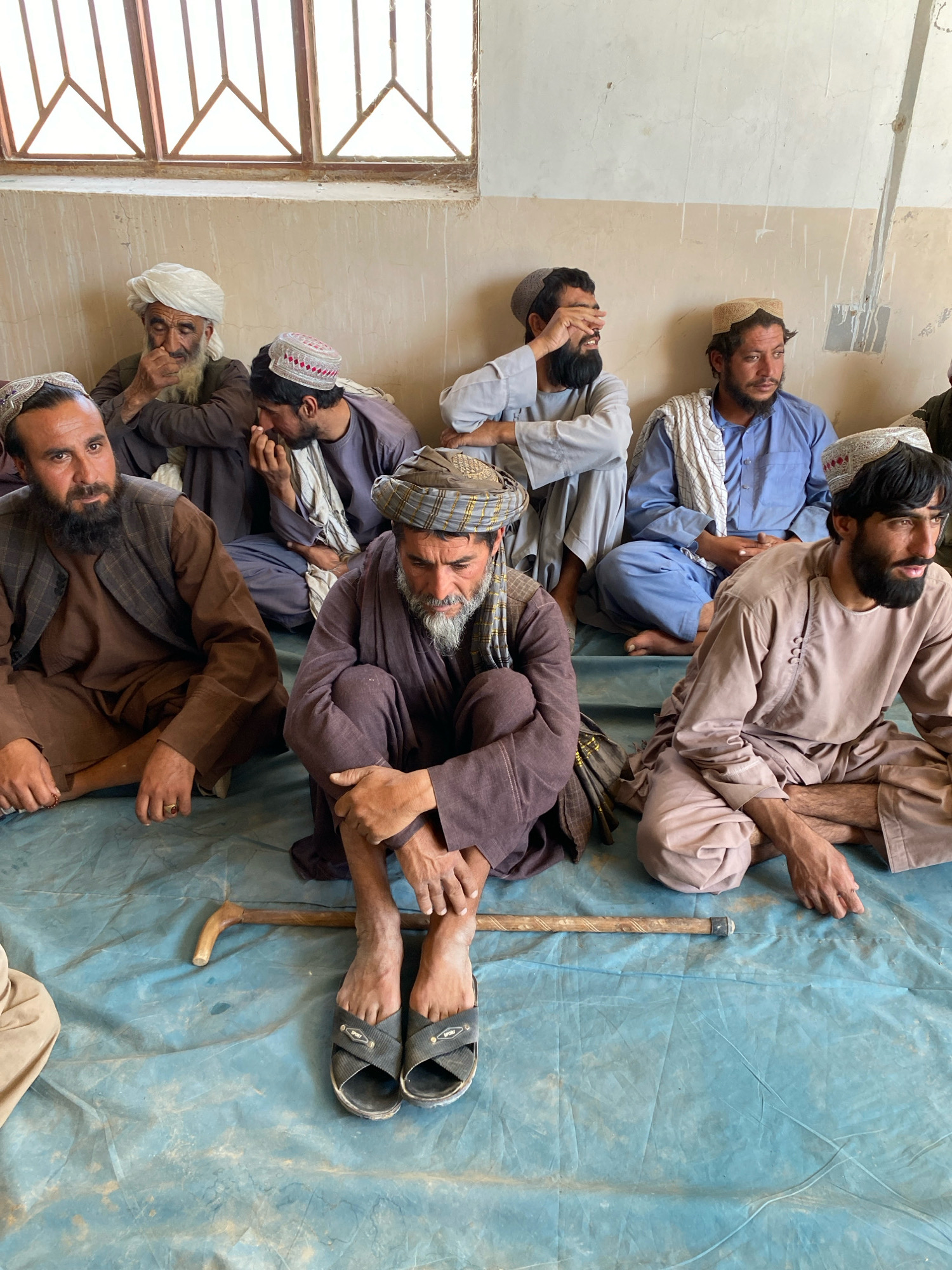 In a room in Pasab, Afghanistan, seven men are seated on a blue tarp. The men are dressed in traditional clothing, including turbans and long robes. Most of them are looking down or away from the camera, with one man covering his eyes. A cane rests on the tarp in front of the seated men. The room appears sparsely furnished, with a window visible in the background. The overall atmosphere seems somber and introspective.